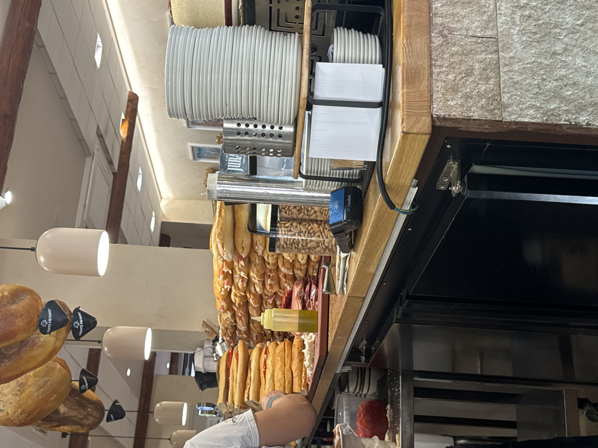 This image depicts a food counter, likely in Murcia, Spain, showcasing a variety of prepared food items. In the foreground, a person, partially visible from the left, is attending to the counter. The counter itself is laden with numerous sandwiches stacked high, some appearing to be filled with meat and condiments like ketchup or tomato sauce. To the right of the sandwiches, a yellow squeeze bottle, possibly containing olive oil, is placed alongside some sliced meats and a container of nuts. Further to the right, a stack of white plates is neatly arranged, suggesting a dining establishment. Above the counter, several cured hams are hanging, adorned with black tags featuring the "MONTE NEVADO" logo. The lighting fixtures are modern, with cylindrical shades emitting a warm glow. The background reveals the interior of the establishment, with visible wooden beams and tiled walls, contributing to a rustic yet functional ambiance.