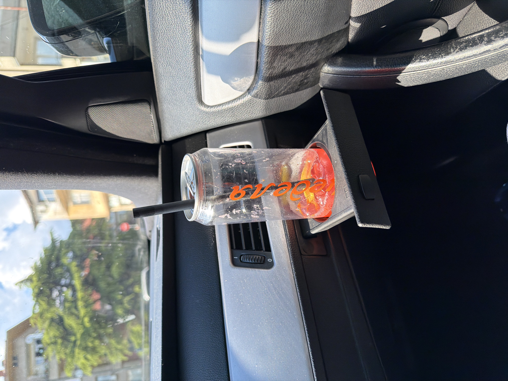 A plastic cup with a black straw rests in a car's cup holder, presumably in Kazanlak, Bulgaria. The cup contains a reddish liquid and a mix of fruit slices. The cup holder is adjacent to a car vent. Outside the car window, a building is visible, along with a large tree. The interior of the car is composed of gray and black plastic.