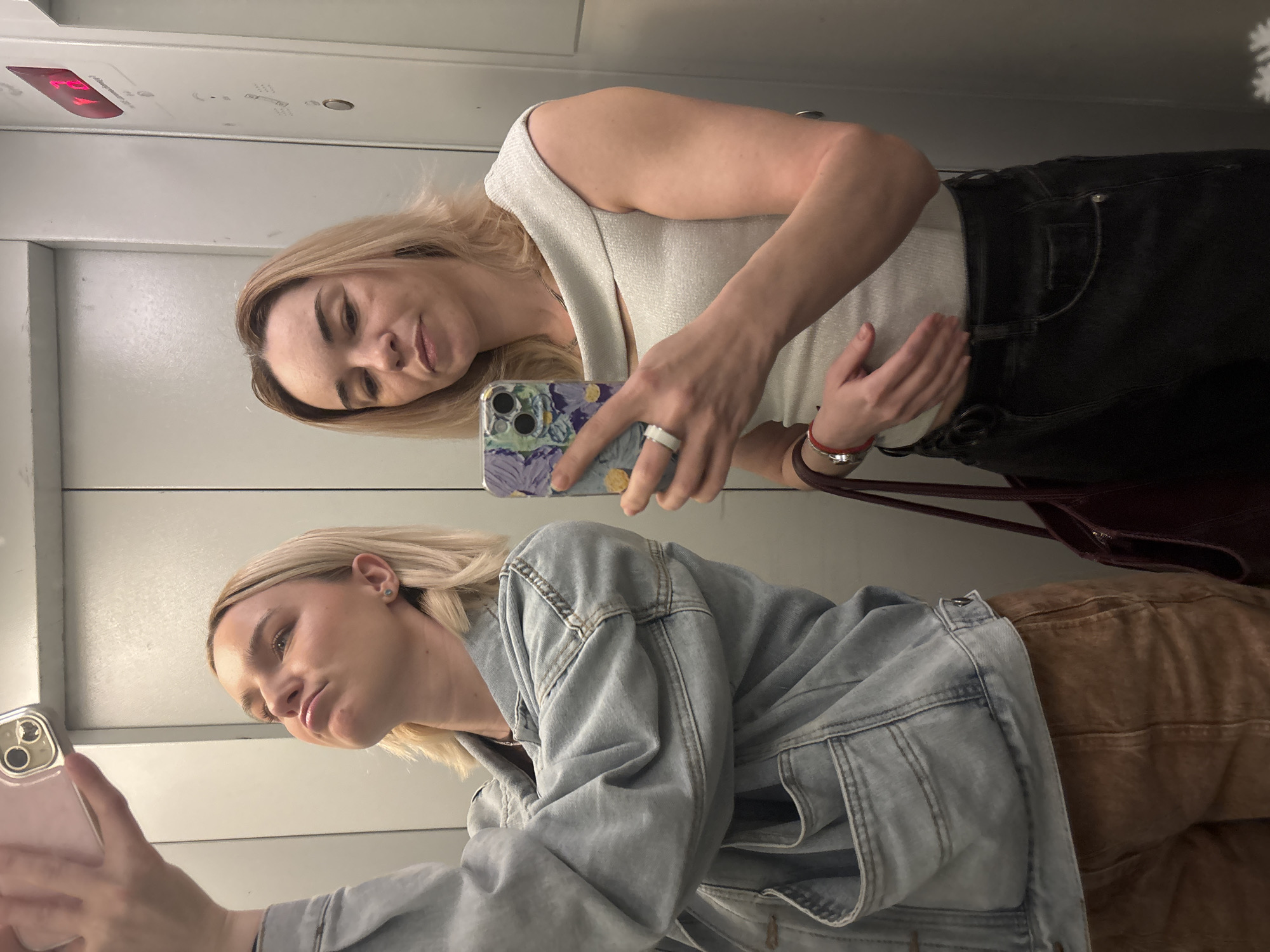 Two light-skinned women are taking a mirror selfie inside an elevator. The elevator's interior is paneled in light gray. The elevator's digital display is visible in the upper left corner. The woman at the top is wearing a light-colored sleeveless top and dark jeans. She is holding a phone with a light-purple decorative case and is wearing a white ring. The woman at the bottom is wearing a light blue denim jacket and tan pants. Her phone has a light pink case. It is unknown if the location of the image is Vsevolozhsk, Russia.