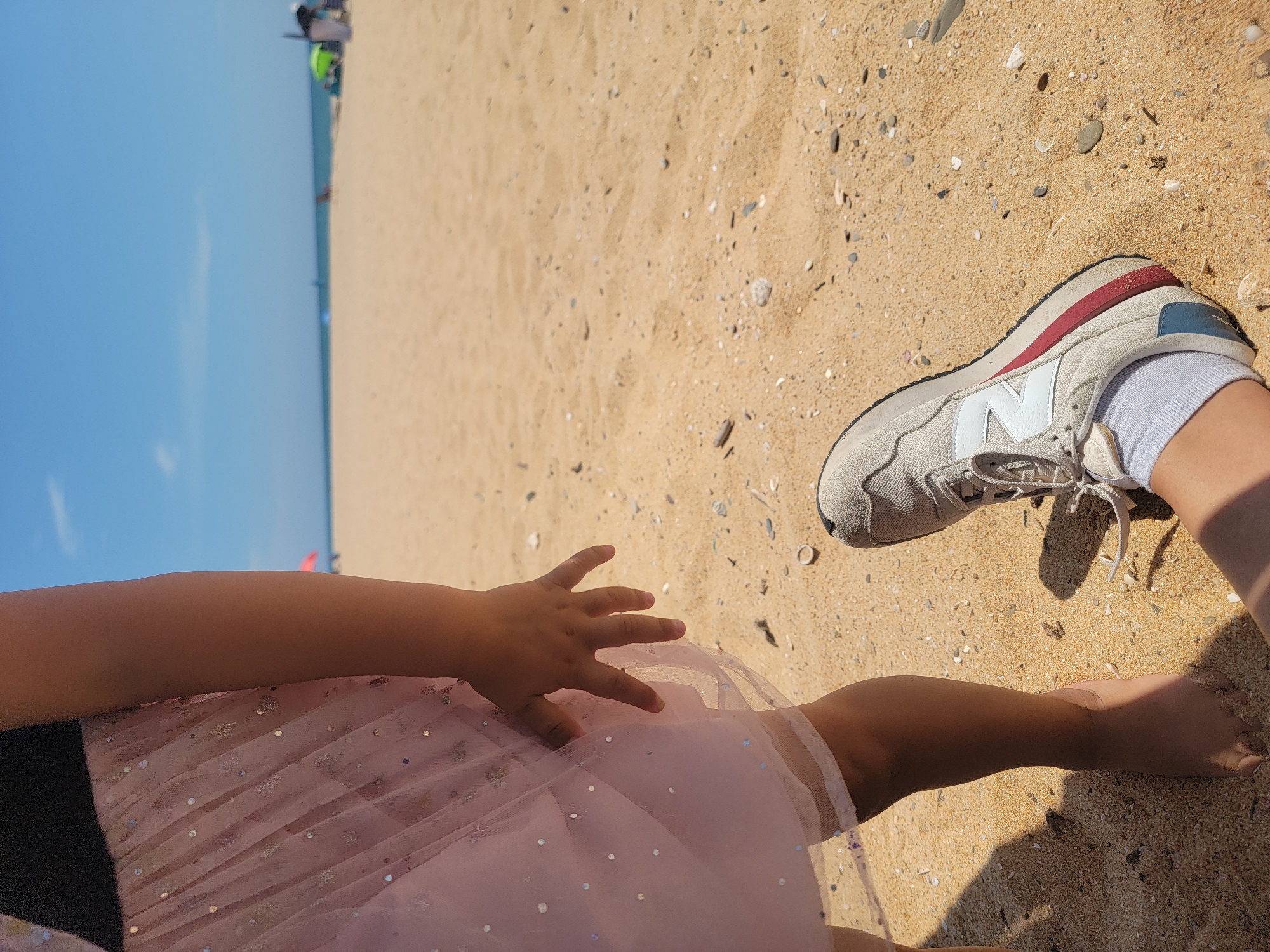 The image shows a close-up of two people's legs and feet on a sandy beach. On the left, a child's bare foot and lower leg are visible, wearing a pink dress with glitter. The child's hand is reaching out near the dress. On the right, an adult's foot is visible, wearing a gray sneaker and white socks. The background depicts a sandy beach with the ocean and a clear blue sky. In the distance, there are beach umbrellas and chairs. The scene is consistent with Quarteira, Portugal, known for its sandy beaches and sunny climate.