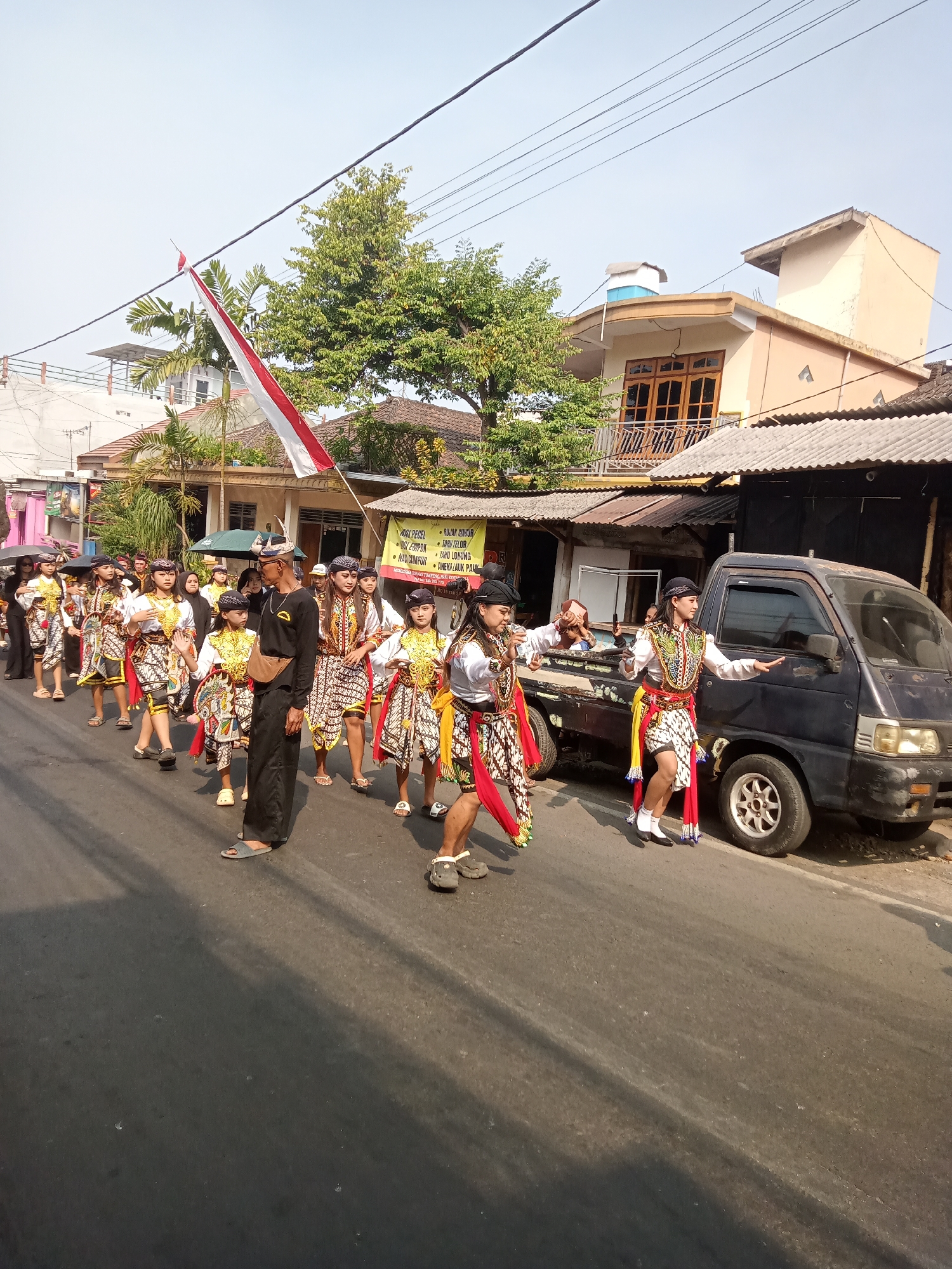 A procession of young people in traditional Javanese attire walks down a paved street in the city of Batu, Indonesia. The participants, some of whom are children and teenagers, wear brightly colored costumes consisting of patterned skirts, white shirts with ornate vests, and headwear. Some individuals hold cultural props. A large red and white flag, representing the Indonesian national flag, is prominently displayed, attached to a pole held aloft by one of the participants.
The procession is moving alongside a dark-colored pickup truck parked on the right side of the street. Buildings line the left side of the street, some with signs displaying text in Indonesian. Trees with green foliage are visible behind the buildings, and electrical wires stretch across the sky above. The scene is illuminated by natural sunlight, indicating it is daytime. The street appears to be a residential or commercial area within Batu, a city known for its cooler climate and agricultural products in East Java, Indonesia. The procession seems to be a cultural event or celebration.
