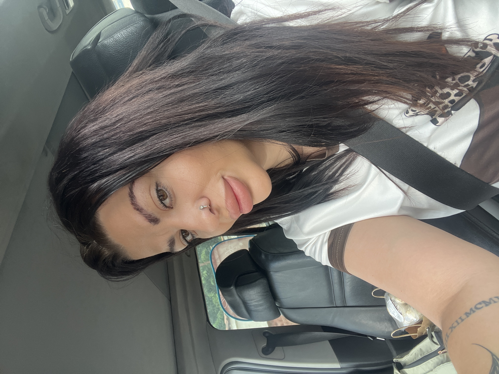 In the city of Kalekovets, Bulgaria, a woman with long, dark hair and a nose ring is seen inside a car. She is wearing a white t-shirt with a brown and leopard print design and a black seatbelt across her chest. The interior of the car, including black leather seats, is visible. The woman is looking towards the camera with a slight smile. Her arm, bearing a Roman numeral tattoo, is also in view.