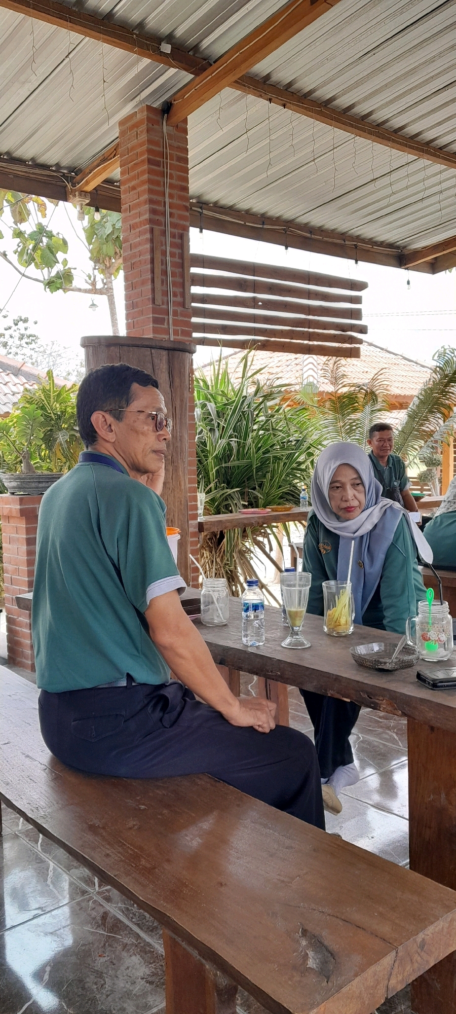 The image shows an outdoor seating area, possibly a cafe or restaurant, located in Sidohasri, Indonesia. A man and a woman are seated at a wooden table. The man, wearing glasses and a green polo shirt, is looking to his left with his hand on his chin. The woman, wearing a hijab and a teal-colored shirt, is looking down. On the table are various drinks in glasses, a bottle of water, and a small dish. In the background, more people are visible, and lush green plants and trees surround the area. The structure above them consists of a metal roof supported by wooden beams and brick pillars. The ground appears to be tiled.