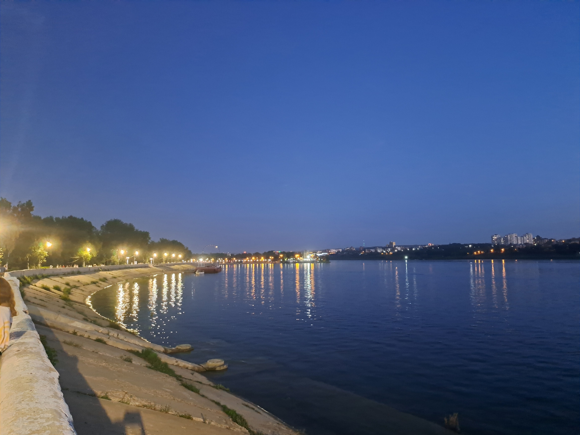 The image depicts a waterfront scene in Irkutsk, Russia, at dusk. The sky is a deep blue, transitioning from a lighter hue near the horizon. A wide river occupies the majority of the frame, with its surface reflecting the lights from the opposite bank and along the embankment.
On the left side of the image, a curved concrete embankment lines the river. Lampposts with bright yellow lights illuminate the walkway along the embankment, casting long reflections on the water. Trees with dark foliage are visible behind the lampposts. A person with brown hair, wearing a horizontally striped top, is partially visible on the far left, leaning on the embankment.
The opposite bank features a cityscape with buildings, some of which are illuminated. A red boat is moored near the embankment on the left side. The overall atmosphere is serene and illuminated by artificial lights against the fading natural light.