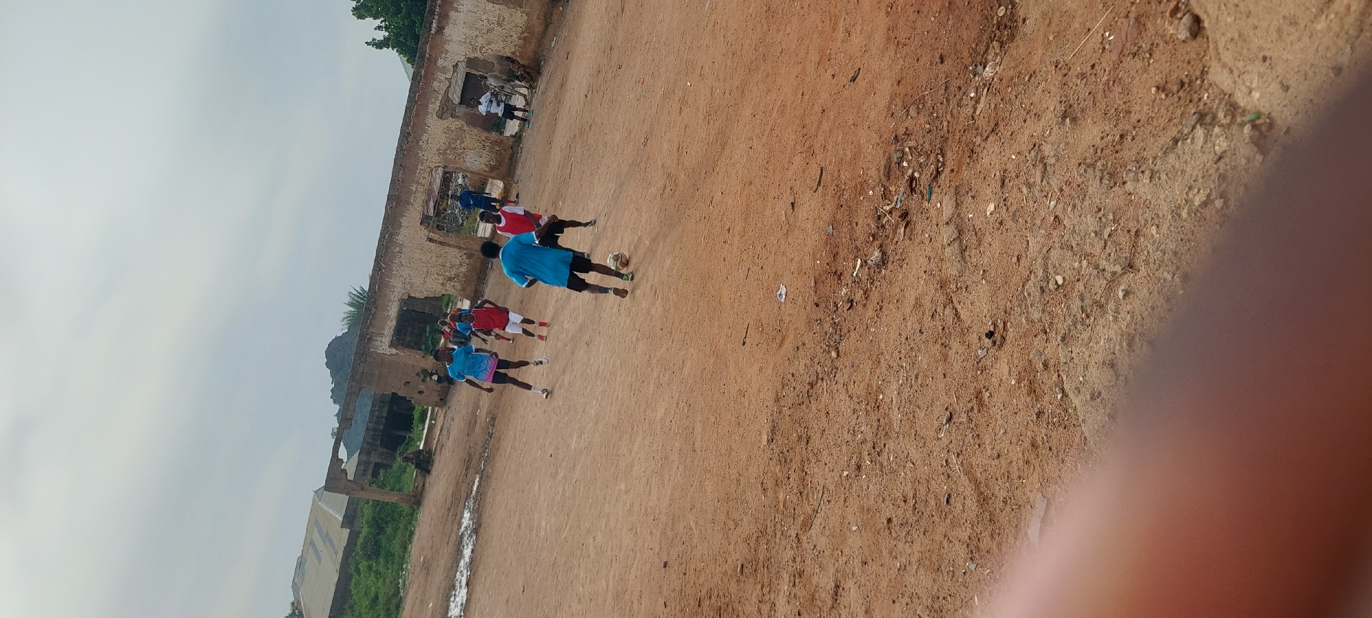 In Bukuru, Nigeria, a group of young men are engaged in a game of soccer on a dirt field. The field is uneven and sandy, with scattered debris. In the background, partially collapsed brick structures with open doorways and roofs are visible, suggesting an abandoned or underdeveloped area. A large, corrugated metal roof structure stands to the left. The sky is overcast, and the overall lighting is diffused. The players are dressed in athletic wear, primarily t-shirts and shorts. One player in a blue t-shirt is dribbling a soccer ball. Another player in a red t-shirt is close by, and others are positioned around the field. A few other individuals appear to be observing the game from the periphery, with one person sitting near the broken structures. The foreground is partially obscured by a blurred, reddish-brown area, likely a thumb or finger in the shot.