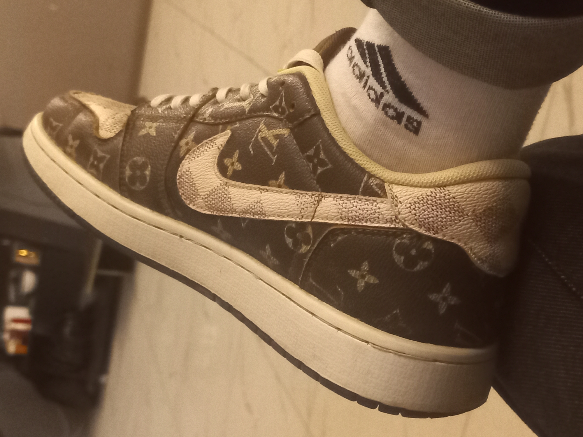 A close-up, low-angle shot captures a person's foot wearing a sneaker and a sock. The sneaker is a low-top design with a brown base adorned with the Louis Vuitton monogram pattern. A white Nike swoosh with a textured, cracked appearance is visible on the side of the shoe. The sole of the sneaker is white. The person is wearing a white sock with black Adidas stripes and the Adidas logo. The background is blurry, suggesting an indoor setting with muted lighting. The overall impression is one of fashion and streetwear.