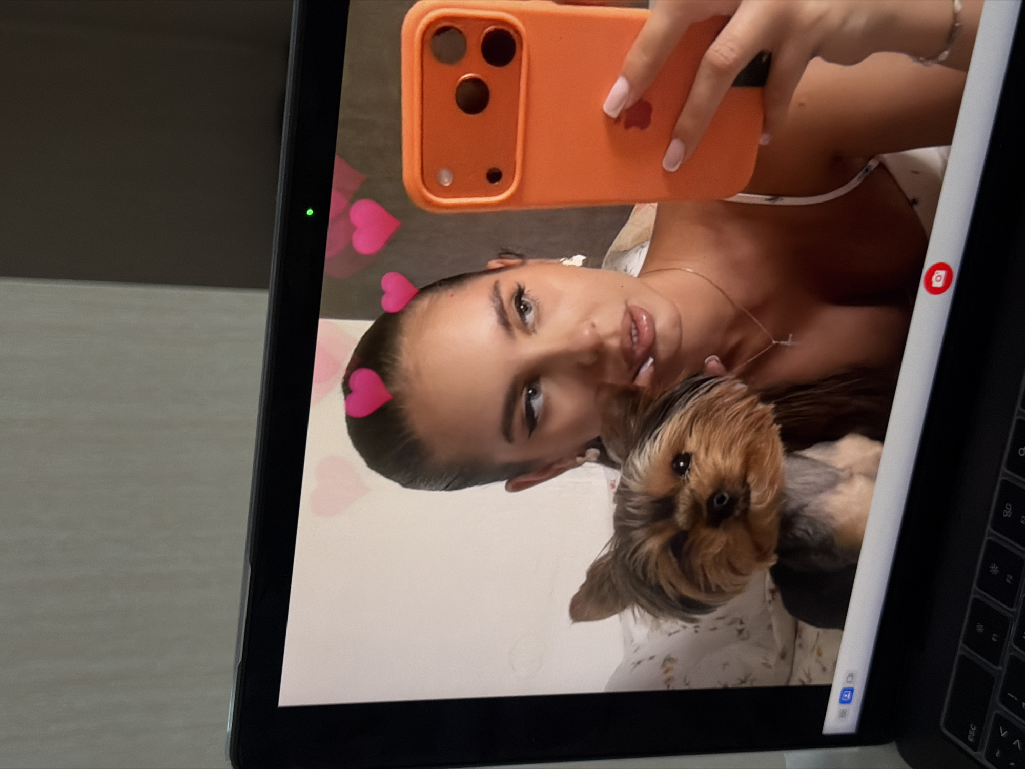 A laptop screen displays a selfie photograph of a woman holding an orange smartphone. The woman has a heart filter over her head and is holding a small dog. The laptop is positioned on a table, visible to the left and bottom. The location is likely indoors but cannot be confirmed, nor can any location in Krasnoyarsk, Russia be discerned.