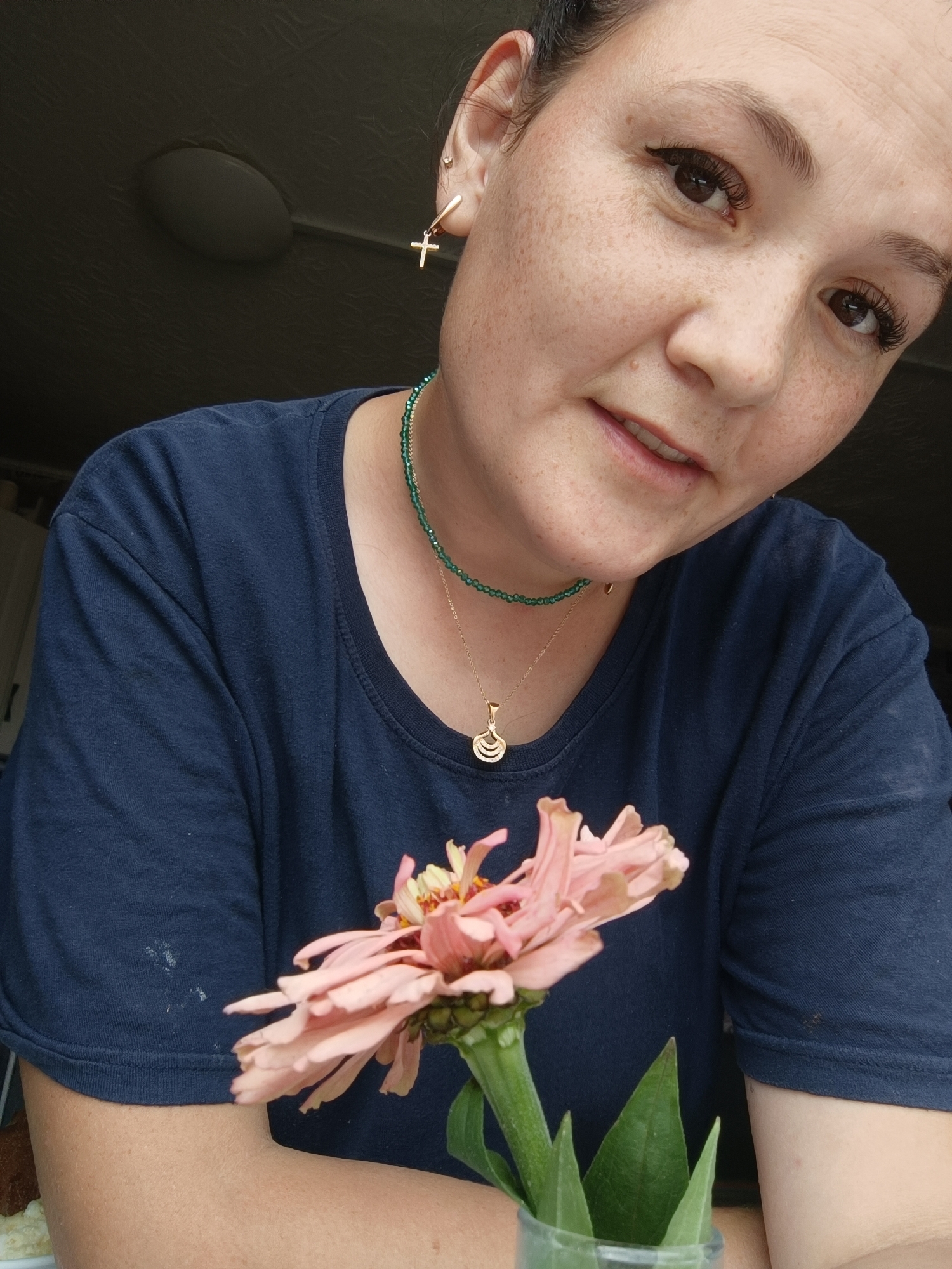 The image is a close-up, waist-up shot of a woman with light skin, brown eyes, and dark hair pulled back. She is wearing a navy blue t-shirt and several pieces of jewelry: a green beaded necklace, a gold pendant necklace, and a gold earring with a cross charm in her left ear. The woman has freckles across her nose and cheeks and is wearing eyeliner. She is holding a pink zinnia flower in front of her chest, with the stem visible in a clear vase. The background is dark and out of focus, with a faint indication of a ceiling fan above and a wall or door to the left. The image does not provide any information about the location being Karsha, Russia.