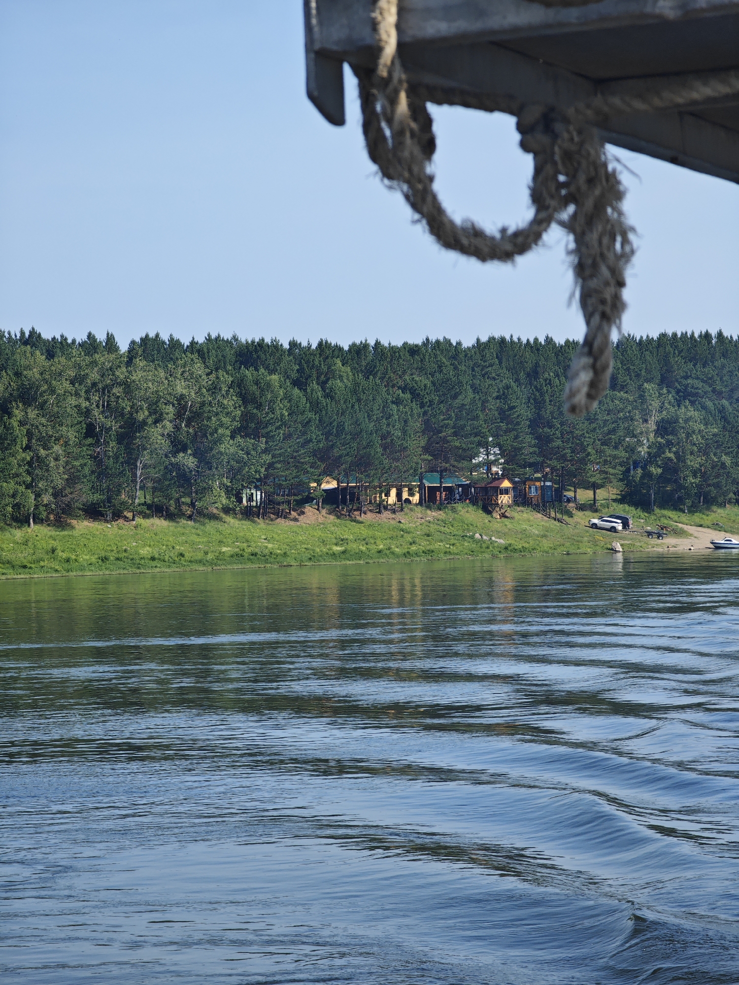 The image captures a serene river scene in Podporozh'e, Russia, on a clear, sunny day. In the foreground, the gentle ripples of the water create a dynamic surface. The middle ground features a lush, green riverbank densely populated with a variety of trees, predominantly evergreens, with a few deciduous trees interspersed. Along the riverbank, a small settlement of colorful buildings, possibly cabins or cottages, is visible amongst the trees. Several vehicles, including a white SUV and a trailer, are parked near the shore, along with a small boat. The background consists of a clear, light blue sky. A thick, weathered rope hangs from the top right of the frame, partially obscuring the sky and the upper edge of the riverbank. The overall impression is one of a peaceful natural setting with signs of human habitation.