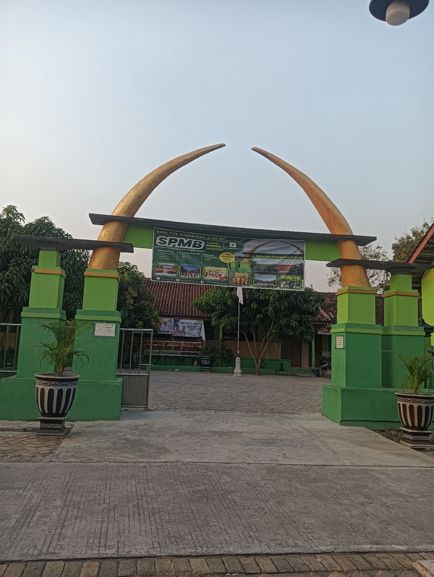This image depicts the entrance gate of SDN Pilangsari 2 Gesi, a primary school located in Dukuh, Indonesia. The gate is characterized by two large, curved golden structures that arch upwards, supported by green pillars. A banner is prominently displayed above the entrance, announcing "SPMB SISTEM PENERIMAAN MURID BARU" (New Student Admission System) for the academic year 2026/2027, with the school's name "SDN PILANGSARI 2 GESI" also visible. The school building with a red tile roof is visible behind the gate, surrounded by trees. Two large decorative pots with plants are placed on either side of the entrance walkway. The ground is paved with concrete and brickwork. The sky is clear and light, suggesting it is either early morning or late afternoon. There are no people visible in the image.