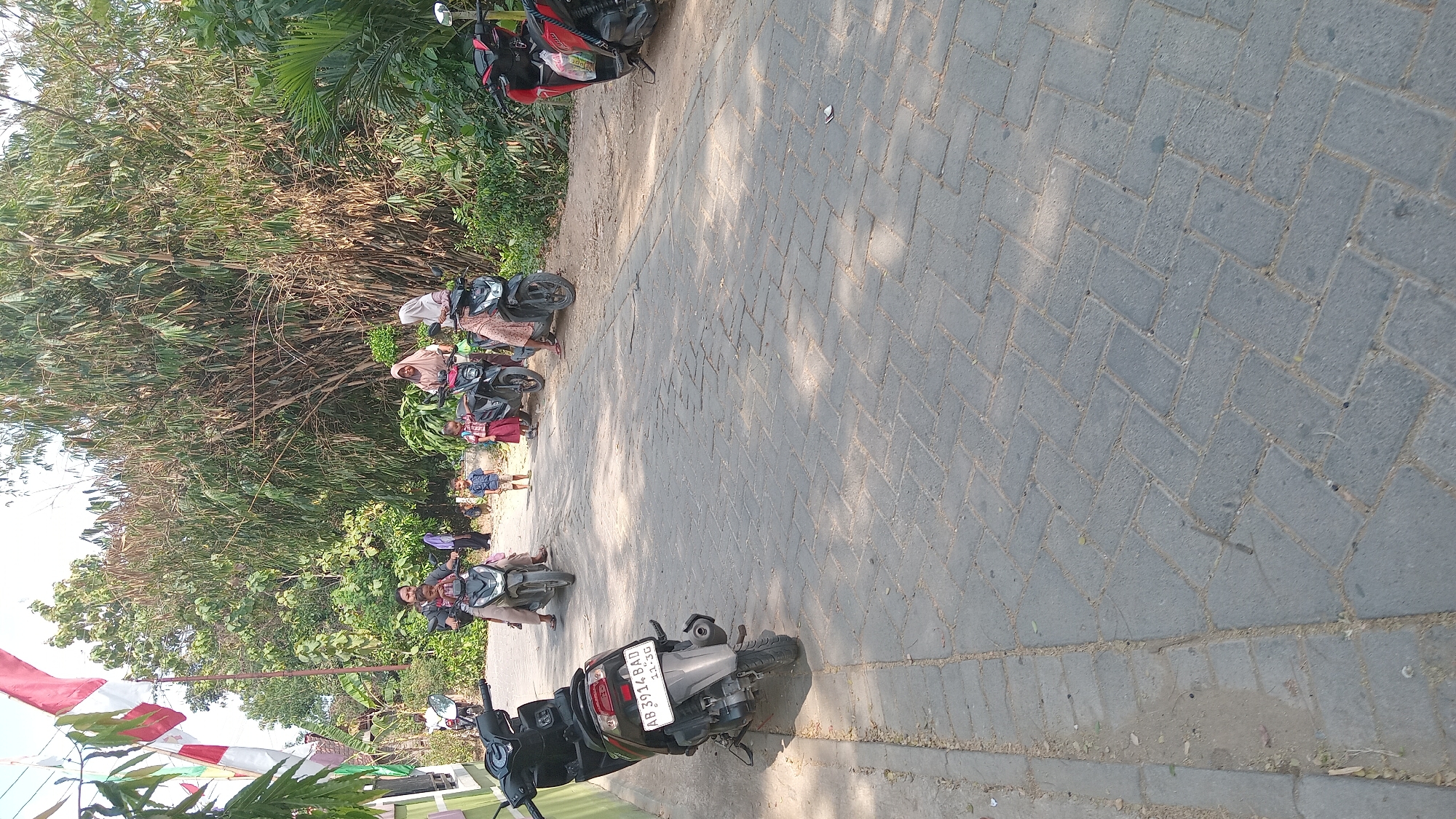 In the city of Kedungwaru, Indonesia, on a paved road with interlocking gray bricks, a group of people are seen near a dense bamboo grove. Several motorcycles are parked and in use. In the foreground to the left, a gray motorcycle with license plate AB 3914 BAD is parked facing the right. Further down the road, a woman is riding a motorcycle with a child on the front. Behind them, two young children, a boy in a blue shirt and shorts and a girl in a red skirt and white top, stand on the side of the road. To the right, two women are on a black motorcycle, and another woman is on a pink and black motorcycle. A red and black motorcycle is parked on the right side of the road. The environment is lush with green foliage and tall bamboo stalks. Flags are visible in the upper left corner, attached to a building.