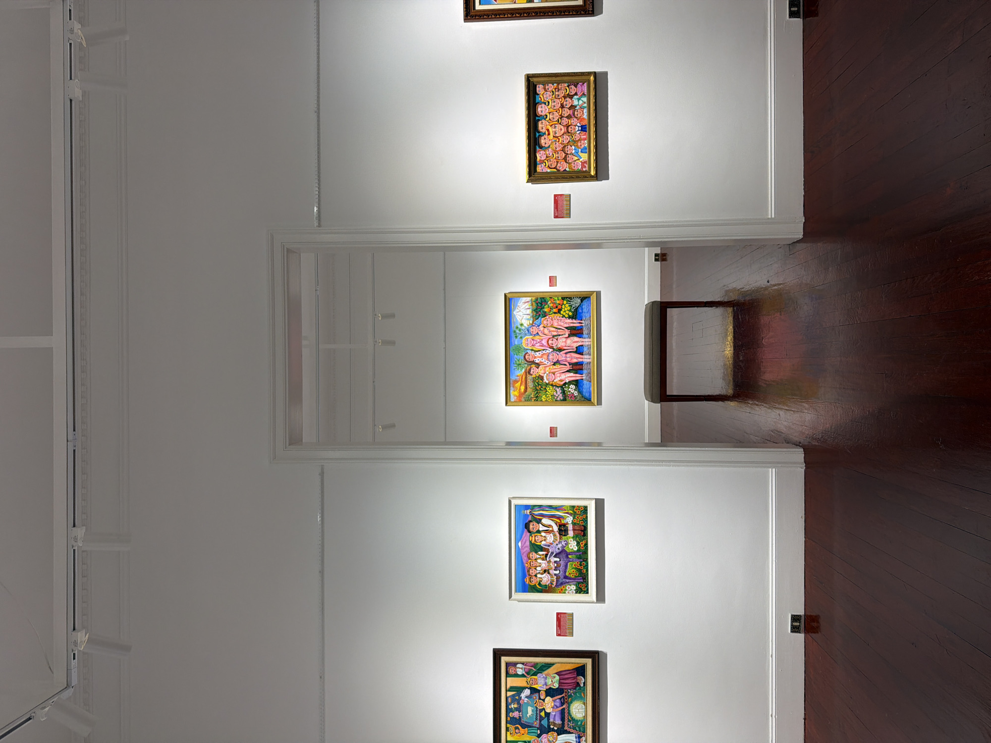 This is a wide shot of an art gallery with polished dark wood floors and white walls. Several paintings are displayed on the walls. On the left, a framed painting depicts a group of people, possibly a family, with a lamb and religious imagery. Next to it, another framed painting shows a family, including a child on a donkey, surrounded by sunflowers with a mountain in the background. In the center, through an open doorway, a bench is placed in front of a painting of a group of nude figures standing in water. To the right of the doorway, another framed painting displays a crowd of people with exaggerated facial features. Further to the right, a sliver of another framed painting is visible. Track lighting is installed on the ceiling.