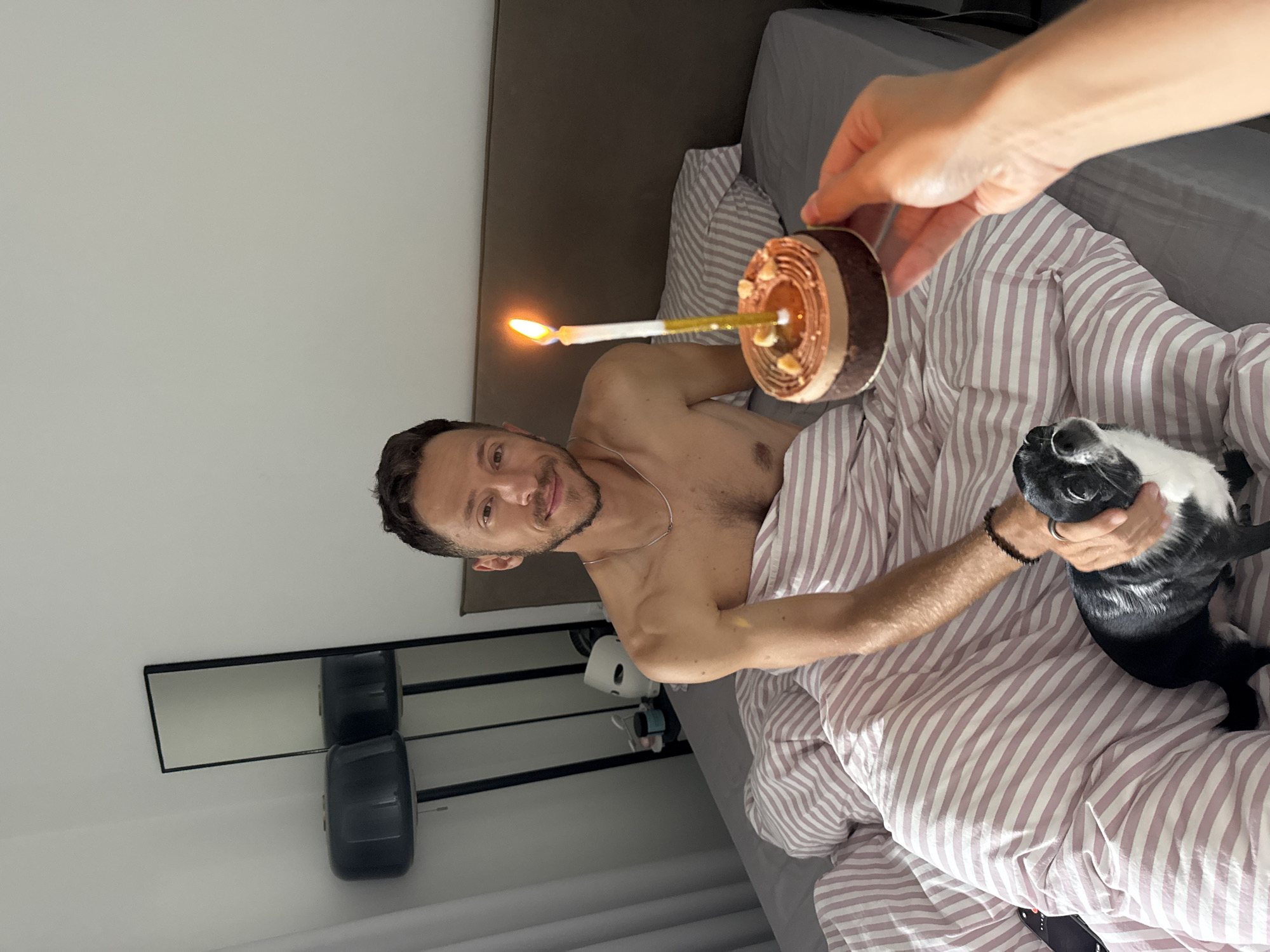 A man is sitting in bed, covered by pink and white striped comforters. He is shirtless and smiling at the camera. In the foreground, a hand is holding a small chocolate cake with a lit candle. A black and white dog is sitting next to the man, looking at the camera. The room appears to be a bedroom with a headboard, a mirror, and lamps visible in the background. The overall scene suggests a birthday celebration.
