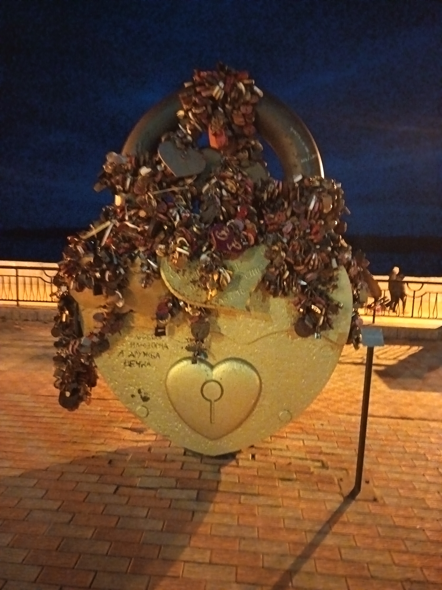 A large, golden lock sculpture stands on a brick walkway in Nizhnevartovsk, Russia. The lock, shaped like a heart, is covered with smaller locks. A metal pole supports the sculpture, and a railing is visible in the background. The sky is dark, suggesting it is night.