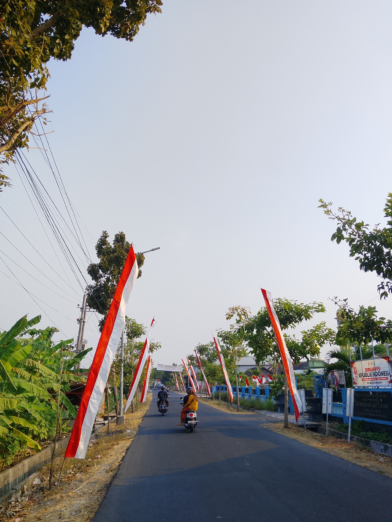 The image depicts a street scene in the city of Ngadrin, Indonesia, likely during a national celebration. Red and white Indonesian flags are prominently displayed along the sides of the road, attached to poles and extending into the sky. Several people are riding on motorcycles, moving down the asphalt road. The environment appears to be somewhat rural or suburban, with trees, vegetation, and utility poles visible. A sign in Indonesian can be seen on the right side of the road, indicating a building or institution. The sky is clear and bright, suggesting it is daytime.