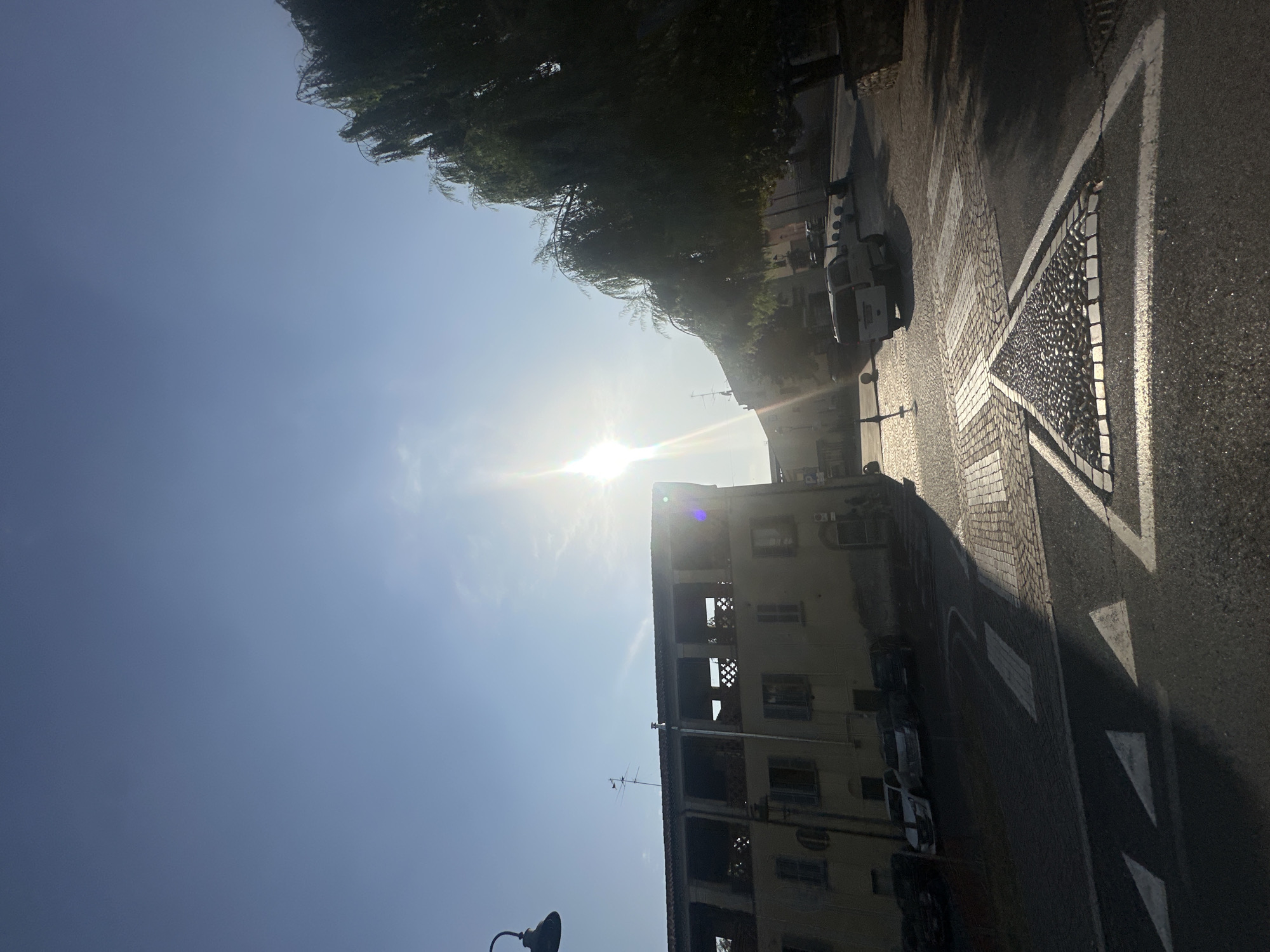 The image depicts a street scene in Chiari, Italy, under a bright, sunny sky. The sun is high, casting strong shadows and creating lens flare, suggesting it is midday or early afternoon. The scene is oriented vertically.
In the foreground, a street with asphalt pavement is visible, marked with white directional arrows and a pedestrian crossing. A mosaic-like patterned area borders the street. The pavement transitions to cobblestones in some areas.
On the left side of the street, a building with multiple floors is partially visible. It appears to be an older building, with some signs of disrepair, such as open window frames and exposed structural elements. An antenna is visible on the rooftop. A streetlamp is also present in the lower left corner.
Several vehicles are parked along the building, including what appears to be a white van or delivery truck.
To the left of the building, a large tree with lush green foliage stretches upwards, partially obscuring the sky. The overall impression is of a quiet street in a European town on a clear, warm day. No people are explicitly visible in the frame, though their presence is implied by the setting. The lack of activity suggests a lull in the day's business.