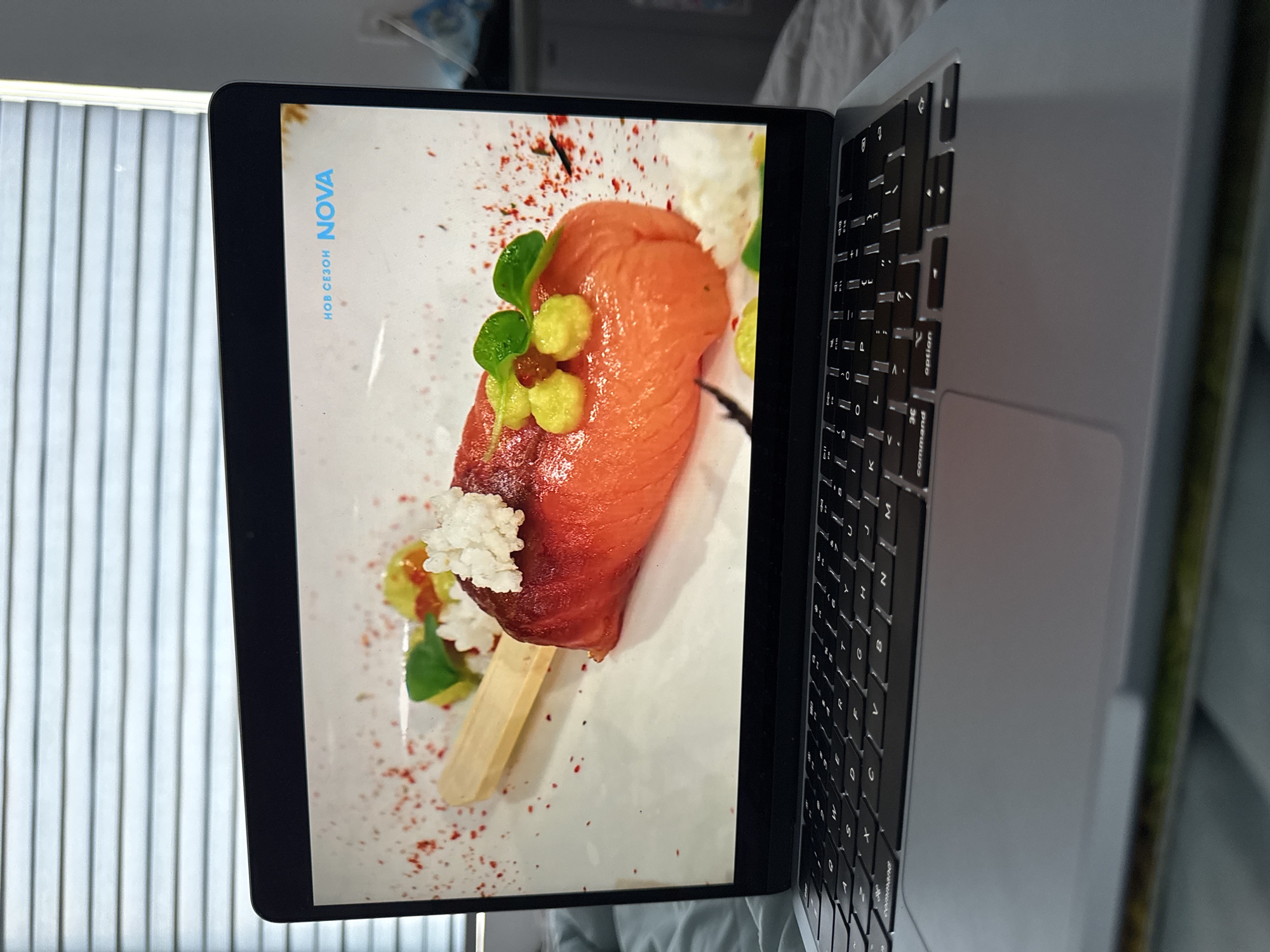 A laptop displays a photo of a dish on a popsicle stick, garnished with what appears to be red pepper flakes, green leaves, and white toppings. "НОВ СЕЗОН NOVA" is printed in blue text on the screen. The laptop rests on a surface with a blurry background. The background appears to be a set of blinds.