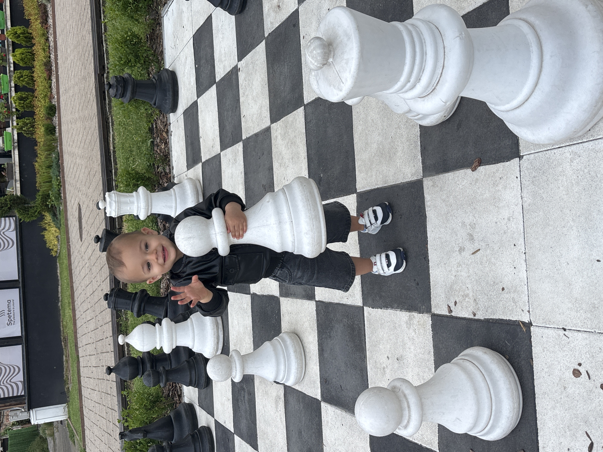 An outdoor scene in Etropole, Bulgaria, features a toddler standing on a large public chess board. The child, appearing male and approximately two years old, is centrally positioned, smiling directly at the viewer with his right hand raised in a greeting gesture. He wears a black leather jacket, dark gray denim shorts, and white sneakers with black and blue accents. In his left arm, he holds an oversized white chess pawn. The black and white checkerboard surface, composed of paved squares, extends across the foreground and midground. Numerous large chess pieces are scattered across the board, including white pawns, a white bishop, and a white queen, alongside black pawns, a black knight, and a black queen. Behind the chess board, a narrow strip of green grass and shrubs separates the playing area from a paved pedestrian walkway. In the background, a dark-colored building with large windows is visible, featuring white signage that reads "Spetema" and "The pleasure is yours!" with a wave logo. Small potted plants line a ledge in front of the building. To the far left, a white utility box is partially visible. The lighting suggests an overcast daytime sky.