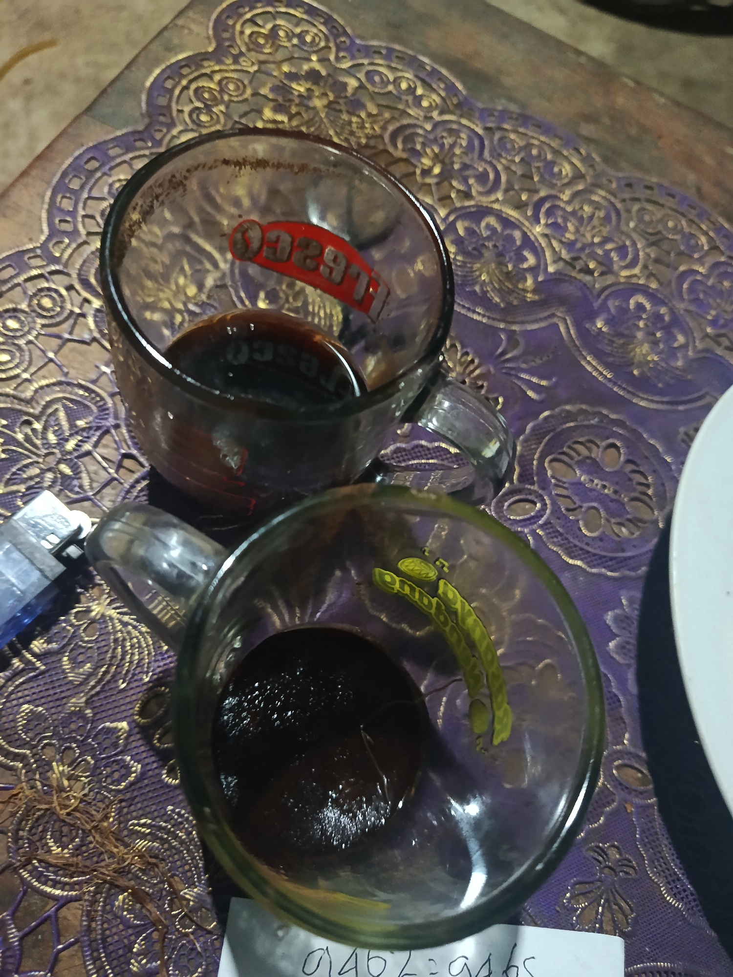 In a close-up, overhead shot, two clear glass mugs with dark liquid are placed on a textured, purple and gold patterned tablecloth. The mug in the upper left has a red and white logo on the inside. To the left of the mugs, a silver and blue lighter lies on the tablecloth. The background consists of a brown wooden surface. A white paper with handwritten markings is partially visible at the bottom of the frame. The scene appears to be indoors, possibly in a home or a small eatery in Karangmojo, Indonesia, given the traditional pattern of the tablecloth.