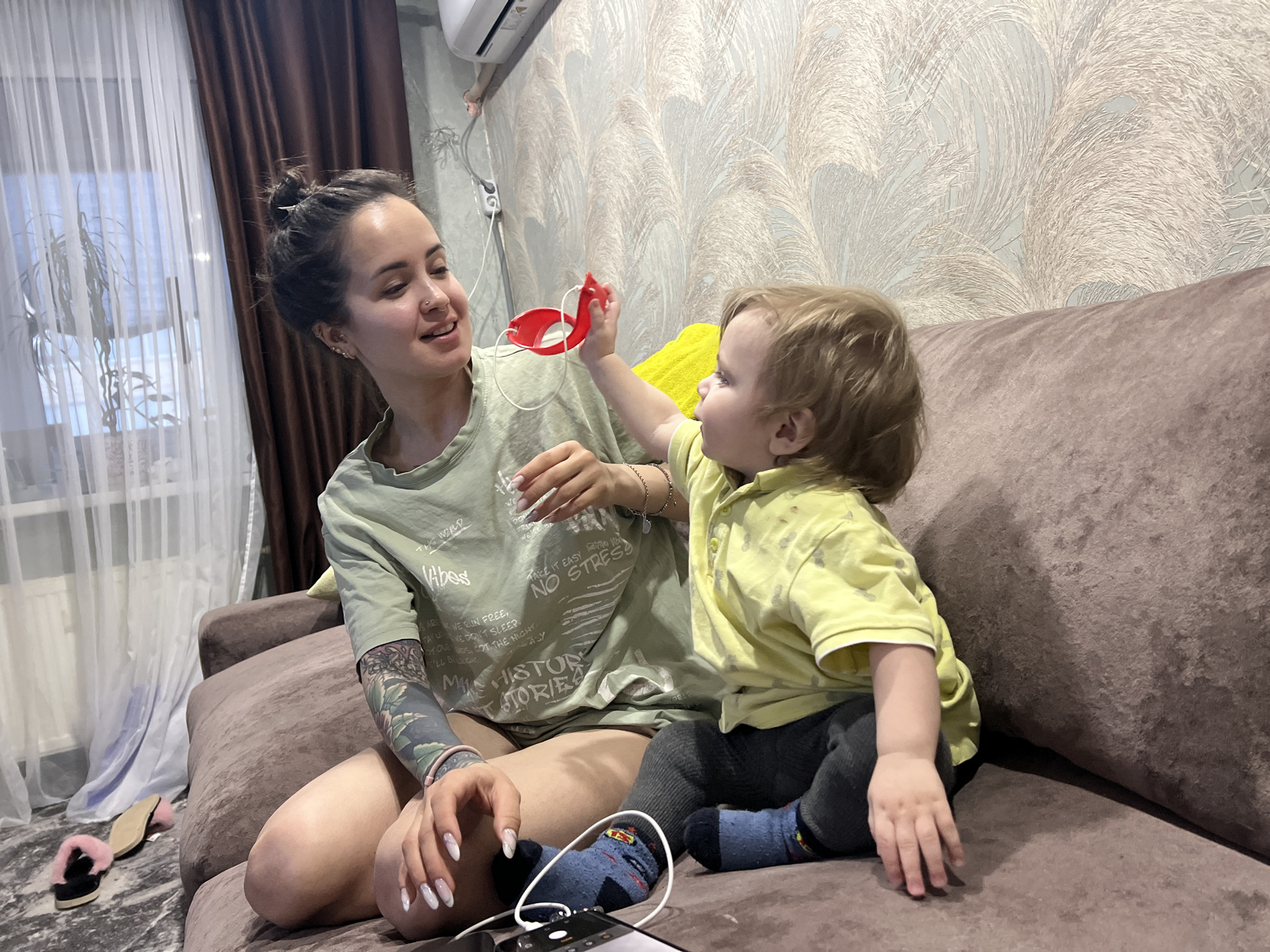 The scene depicts an indoor residential setting in Noviy, Russia, featuring two individuals on a brown upholstered sofa. On the left, an adult female with dark hair tied in a bun, visible nose and ear piercings, and a tattoo sleeve on her left arm, wears a light green t-shirt with text graphics. Her nails are long with white tips. She is looking towards the child, smiling slightly. On the right, a young child with light-colored hair, wearing a yellow polo shirt, dark pants, and blue patterned socks, is holding a red object with a white cord and extending it towards the adult. A black smartphone with a white charging cable is positioned on the sofa between the adult's legs and the child's feet. To the far left, two pink fluffy slippers are on a dark patterned floor near the sofa. Behind the sofa, a wall is covered with textured wallpaper featuring a light, feathery pattern. A window with white sheer curtains and dark brown opaque curtains is visible on the left, with a houseplant partially obscured behind the sheer curtain. An air conditioning unit is mounted on the wall above the window.
