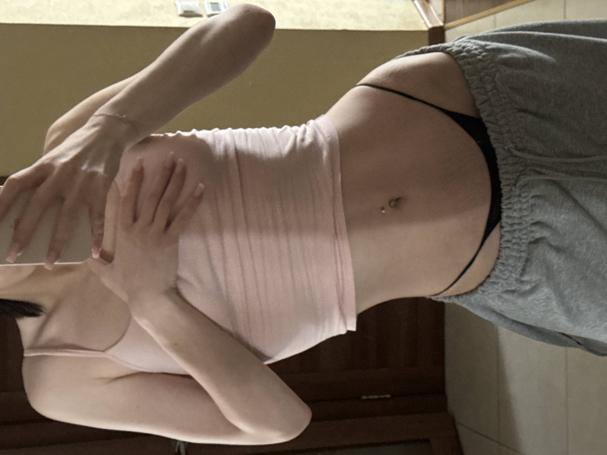 A person is standing indoors, holding a phone to take a mirror selfie. They are wearing a pink tank top, black thong, and grey sweatpants. The background appears to be a room with wooden doors and a yellow wall. There is no indication of the location being in Krasnogorsk, Russia.