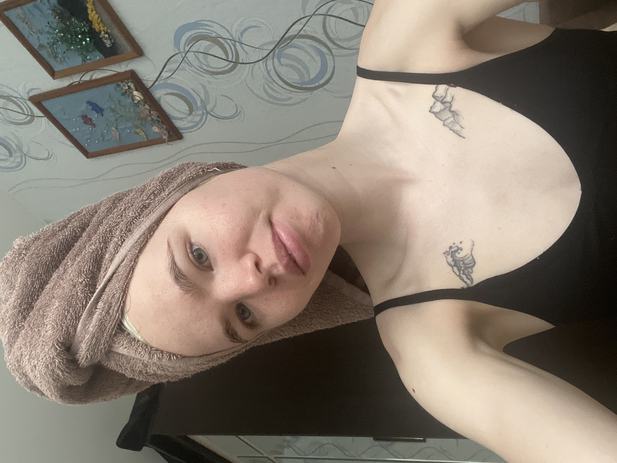 A young fair-skinned person with a towel wrapped around their head is facing the camera. They are wearing a black top. They have two tattoos on their upper chest, one on each side. The background includes a wall with a swirling blue pattern and two framed decorations. The location of Chaikovsky, Russia, cannot be determined from the image.