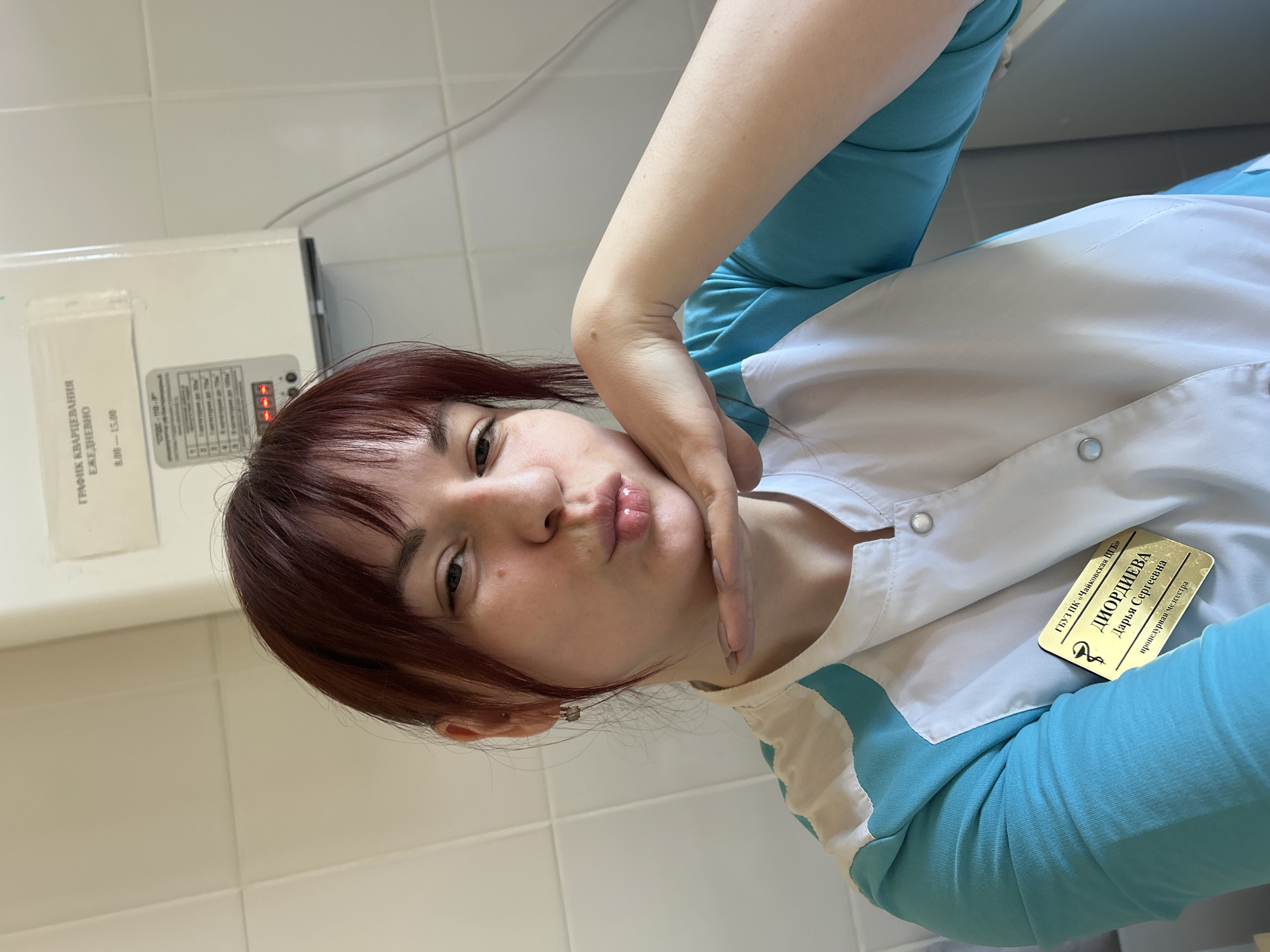The image shows a woman with reddish-brown hair wearing a white and teal uniform, suggesting she is a medical professional. She is posing with her hand near her face. A name tag on her uniform reads "DIORDIEVA Darya Sergeevna," with the title "Procedural Nurse." In the background is a schedule for "QUARTZ TREATMENT" which is "DAILY 8.00-15.00." The name tag also indicates "GBUZ PK 'Tchaikovsky SB'," which is likely a reference to a hospital or medical facility located in the city of Tchaikovsky, Perm Krai, Russia. The image appears to be taken indoors, possibly in a hospital setting, indicated by the tiled walls.