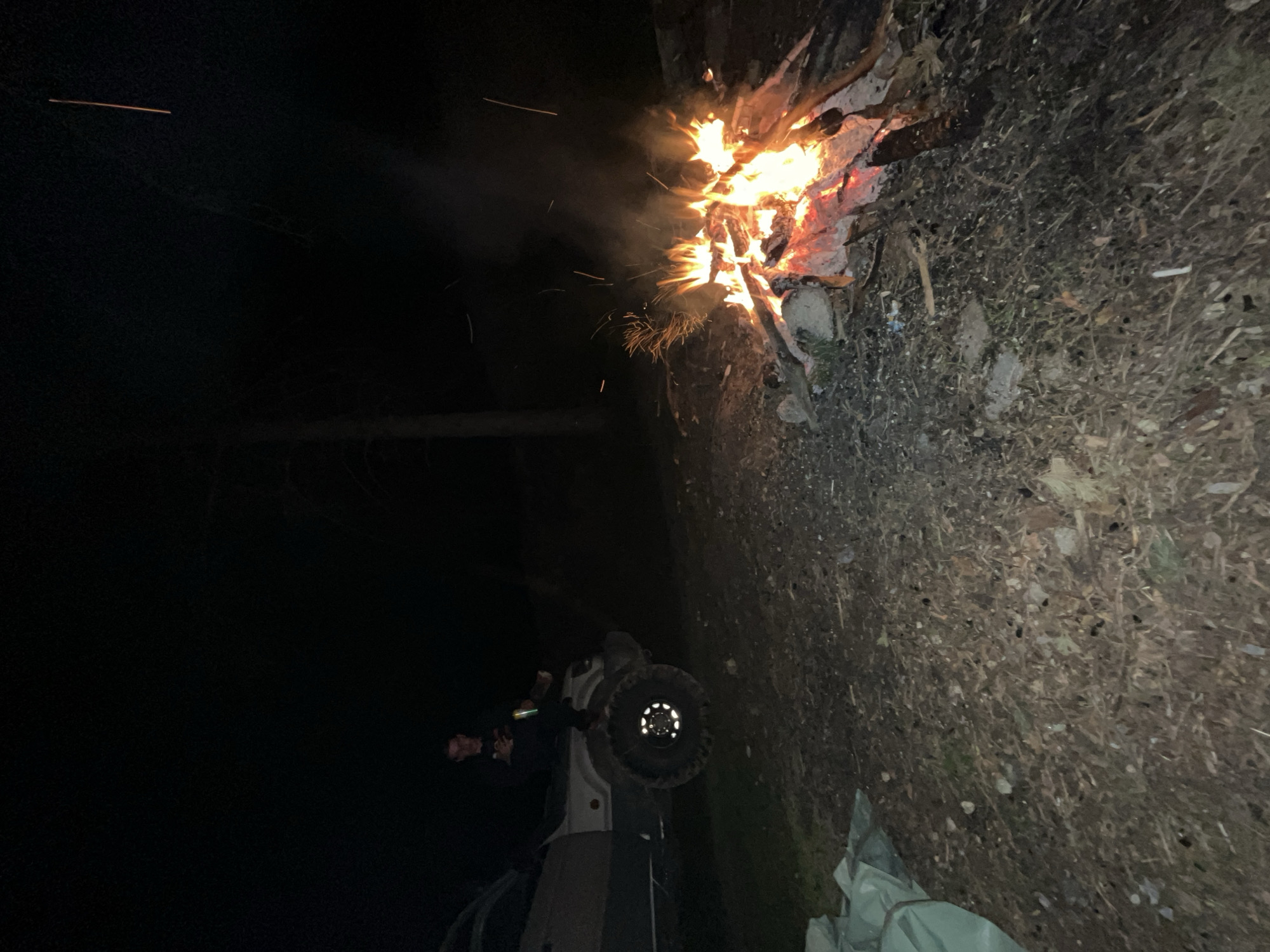 The image shows a night scene in a dark, possibly outdoor location. A campfire is burning brightly on the right side of the frame, casting light on the surrounding area. The ground appears to be covered in dry leaves and debris. In the lower part of the image, there is a partially visible object that resembles a tire and possibly a portion of a vehicle, with a person nearby holding a lit object. The left side of the image is obscured by darkness, possibly indicating a wooded area or a lack of lighting. The image does not include specific details that can confirm it is located in Karavelovo, Bulgaria.