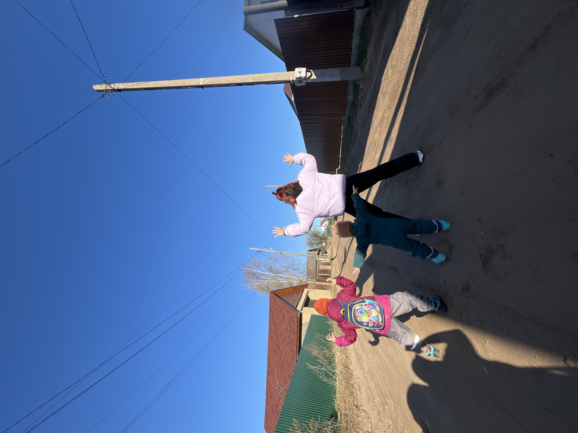 This image, taken in Chaikovsky, Russia, captures a bright, sunny day with three individuals on a dirt road. The scene is rotated 90 degrees counter-clockwise from its natural orientation.
The subjects include an adult and two children, all appearing to be playing or posing with arms outstretched. The adult, wearing a lilac puffer jacket, black pants, and a black face mask, stands further back. One child, dressed in a dark blue/green jacket and pants with light blue shoes, is positioned in the middle. The second child, in the foreground, wears a pink jacket, grey pants, and an orange hat, carrying a vibrant backpack adorned with colorful cartoon characters. Their long shadows stretch across the dirt road, indicating either morning or late afternoon.
The setting appears to be a rural or semi-rural area. A concrete utility pole with multiple power lines extending from it dominates the left side of the frame against a clear, deep blue sky. A small meter or device is attached to the pole. In the background, various buildings are visible, including one with a brown corrugated metal fence and another with a reddish-brown tiled roof and a green corrugated metal fence. Bare trees are scattered between the structures. The ground is a light-colored dirt road with tire tracks.
The clear sky and strong shadows suggest bright, sunny weather, possibly cool given the jackets worn by the individuals. There is no discernible text visible on any objects or clothing, beyond the general design on the child's backpack.