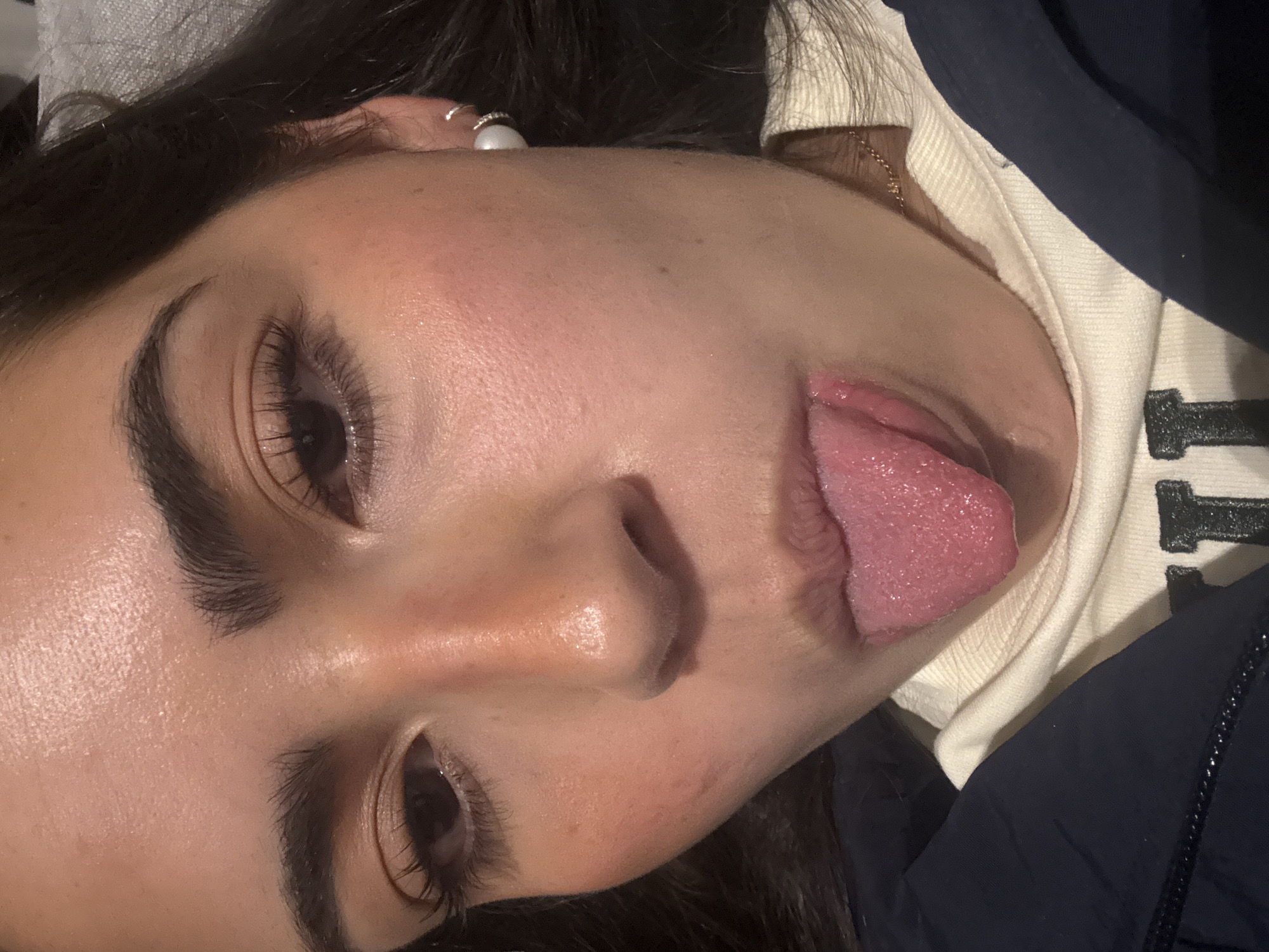 A close-up, slightly tilted headshot of a person with fair skin, dark hair, and dark eyebrows. The person's eyes are open, and their tongue is extended out of their mouth. They appear to be wearing a white shirt with dark lettering, possibly a sweatshirt, and a dark-colored jacket. A small, white pearl earring is visible in their left ear. The background is blurry and undefined. The location in Mexico City, Mexico is not discernible from the image.