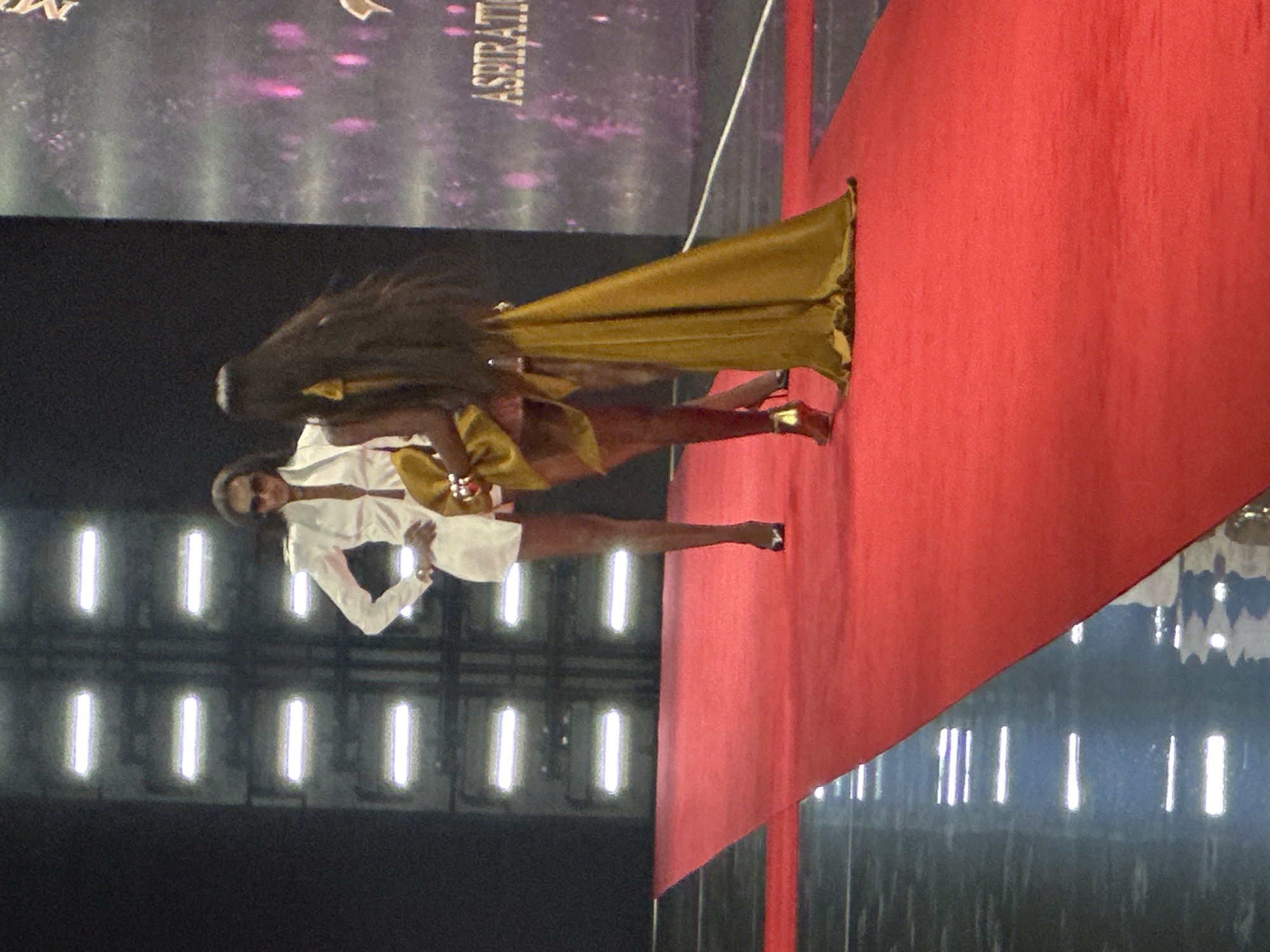 Two women are walking on a red carpet, possibly during a fashion show or event. The woman on the left is wearing a white blazer dress and sunglasses, with her left hand on her hip. The woman on the right is wearing a voluminous gold dress and has long dark hair. The background features a dark wall with illuminated horizontal panels and a screen displaying text that includes "ASPIRATION". The scene appears to be indoors. The provided context does not mention Nowy Sącz, Poland.