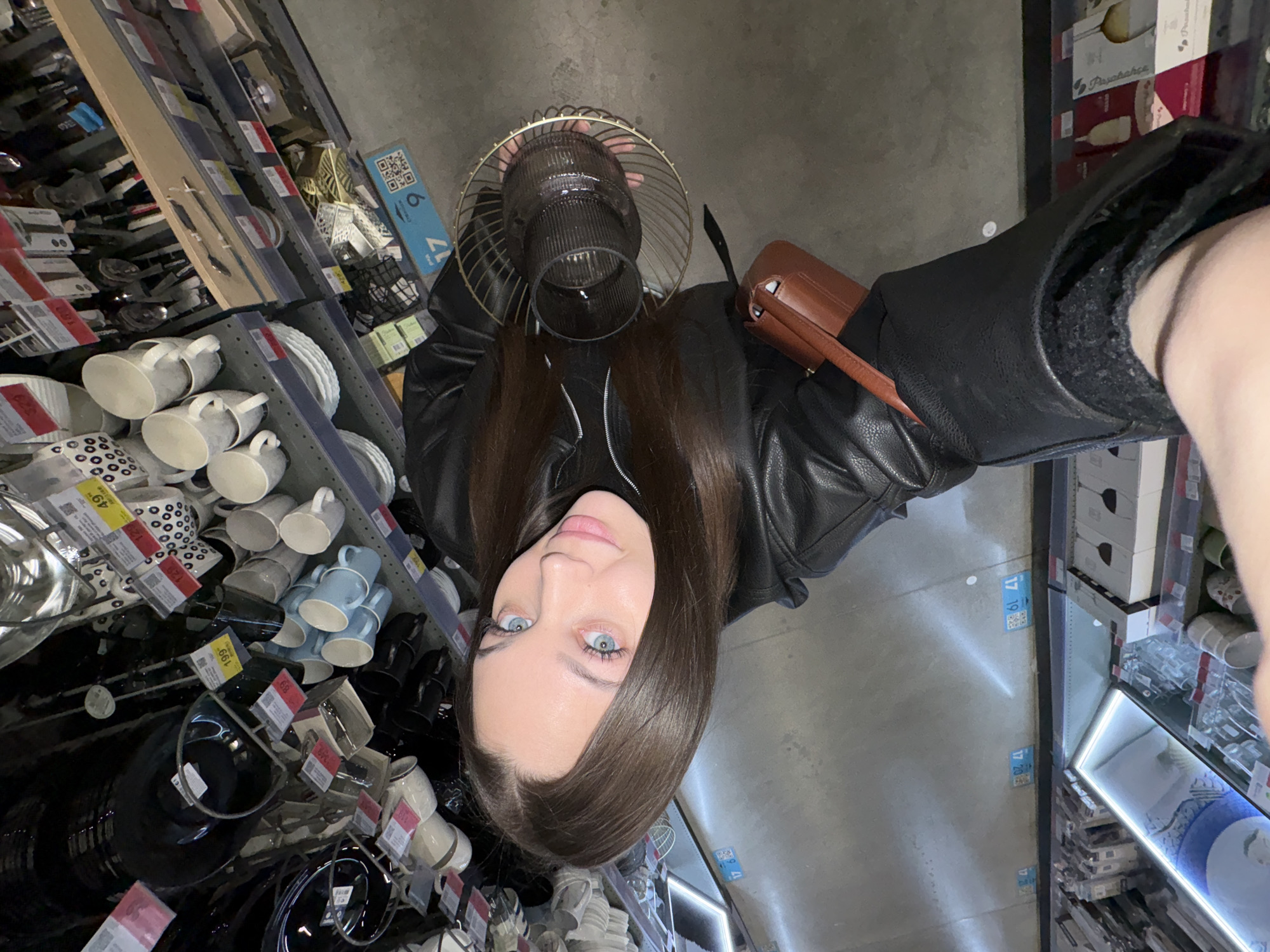 The image is a selfie taken from a top-down perspective, rotated 90 degrees counter-clockwise, showing a young woman in a retail store setting in Yekaterinburg, Russia.
The primary subject is a woman with long, straight dark brown hair and striking blue eyes, looking directly at the camera with a neutral to slight smile. She has fair skin and is wearing a black leather jacket over a dark-colored top. A brown leather-like shoulder bag is visible on her right side. Her left hand is holding a decorative glass object with a gold-colored wire cage design, possibly a lamp shade or a vase.
The scene is an indoor retail space, likely a homeware or kitchenware section of a supermarket or department store. Numerous shelves filled with merchandise are visible around her. These include stacks of white, light blue, black, and patterned ceramic mugs, plates, and bowls. Other items like cutlery sets, various glassware (e.g., wine glasses), and other kitchen and home decor items are also on display. The store has a grey floor and typical artificial retail lighting.
Notable details include several price tags attached to items, featuring numbers like "49", "69", "129", and "199" (likely in Russian Rubles). On the floor, blue adhesive markers with QR codes and numbers are visible, indicating sections or aisles. One marker clearly shows "17 6", another "17 19", and a third "17 20". Some product packaging in the background shows the brand name "Pasabahce", a well-known glassware manufacturer. The overall impression is a casual shopping excursion.