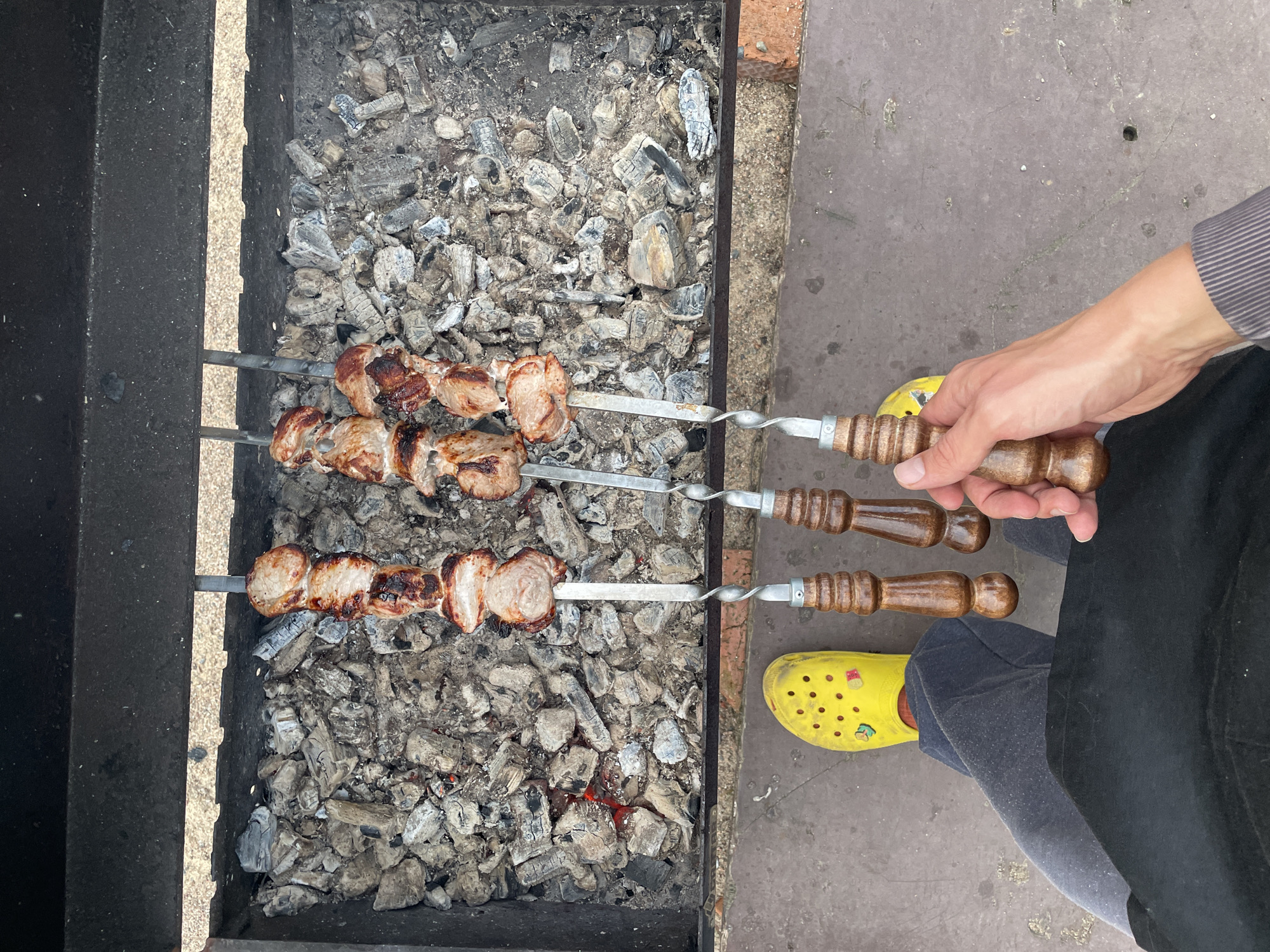 A man is cooking shish kebab over a charcoal grill. The grill is made of metal and contains hot coals. Three metal skewers with meat are visible on the grill, the meat on two of the skewers appearing fully cooked and the meat on the other skewer less so. The man is wearing a gray long-sleeve shirt, black apron, gray pants, and yellow Crocs. The scene appears to be an outdoor area in Sestroretsk, Russia.