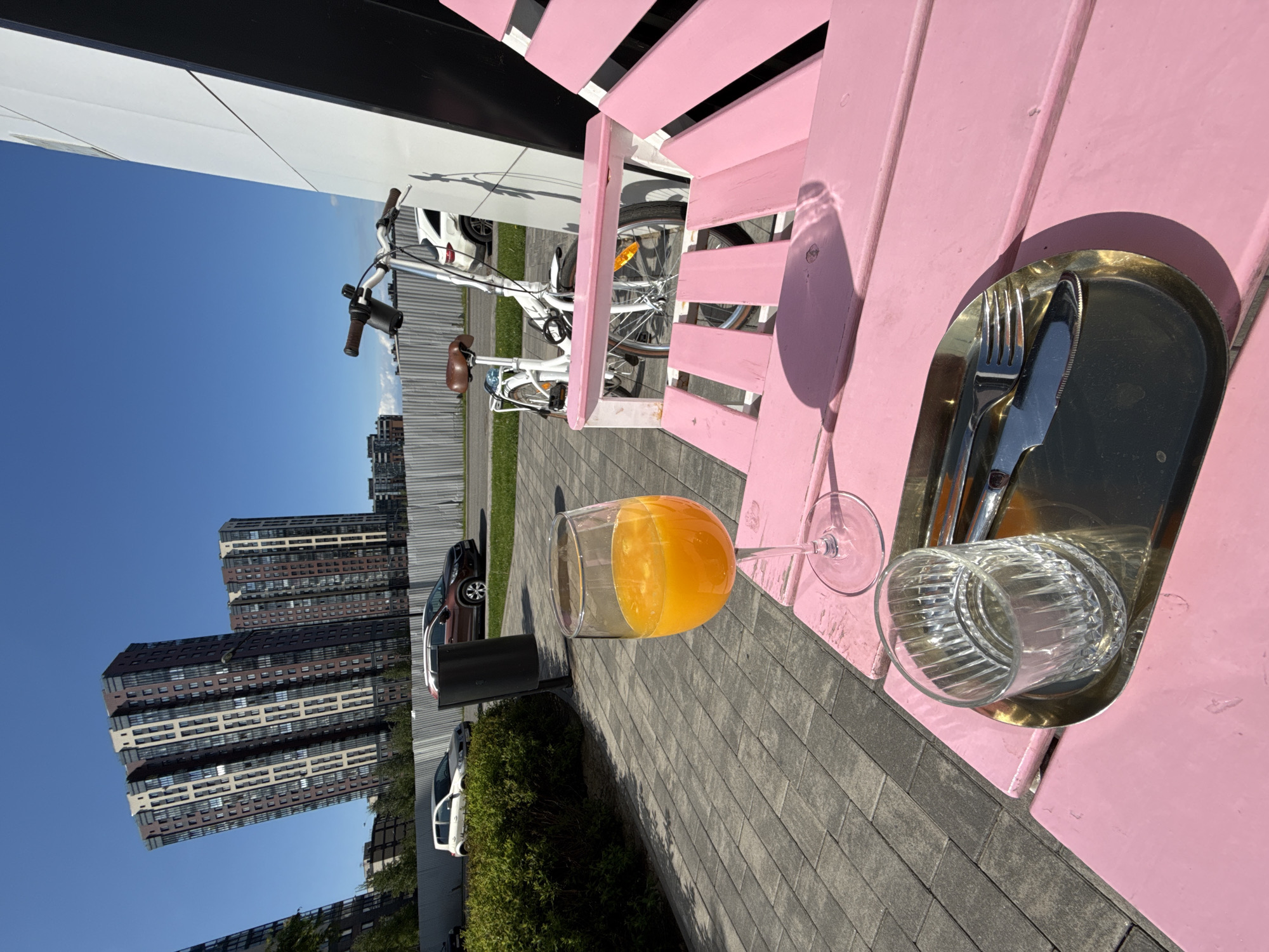 This bright outdoor image captures a vibrant pink wooden bench or table in the foreground, bathed in strong sunlight. On the table, a wine glass filled with orange liquid, presumably orange juice, stands next to an empty clear glass. A small, oval, brass-colored tray holds a fork and a knife, suggesting a place setting for a meal or refreshment. Notable shadows are cast by the objects and the table itself, indicating a sunny day, likely morning or early afternoon.
Behind the pink seating, a white bicycle with brown handlebar grips and seat is parked. The ground beneath is paved with gray rectangular blocks. In the mid-ground, a row of green bushes and several parked cars, including a dark brown and a white vehicle, are visible. The background features a clear blue sky and a cluster of tall, modern multi-story buildings. These urban structures display a pattern of dark and light-colored paneling with numerous windows. The overall scene suggests a pleasant and relaxed outdoor setting in an urban environment. There is no visible text in the image.
