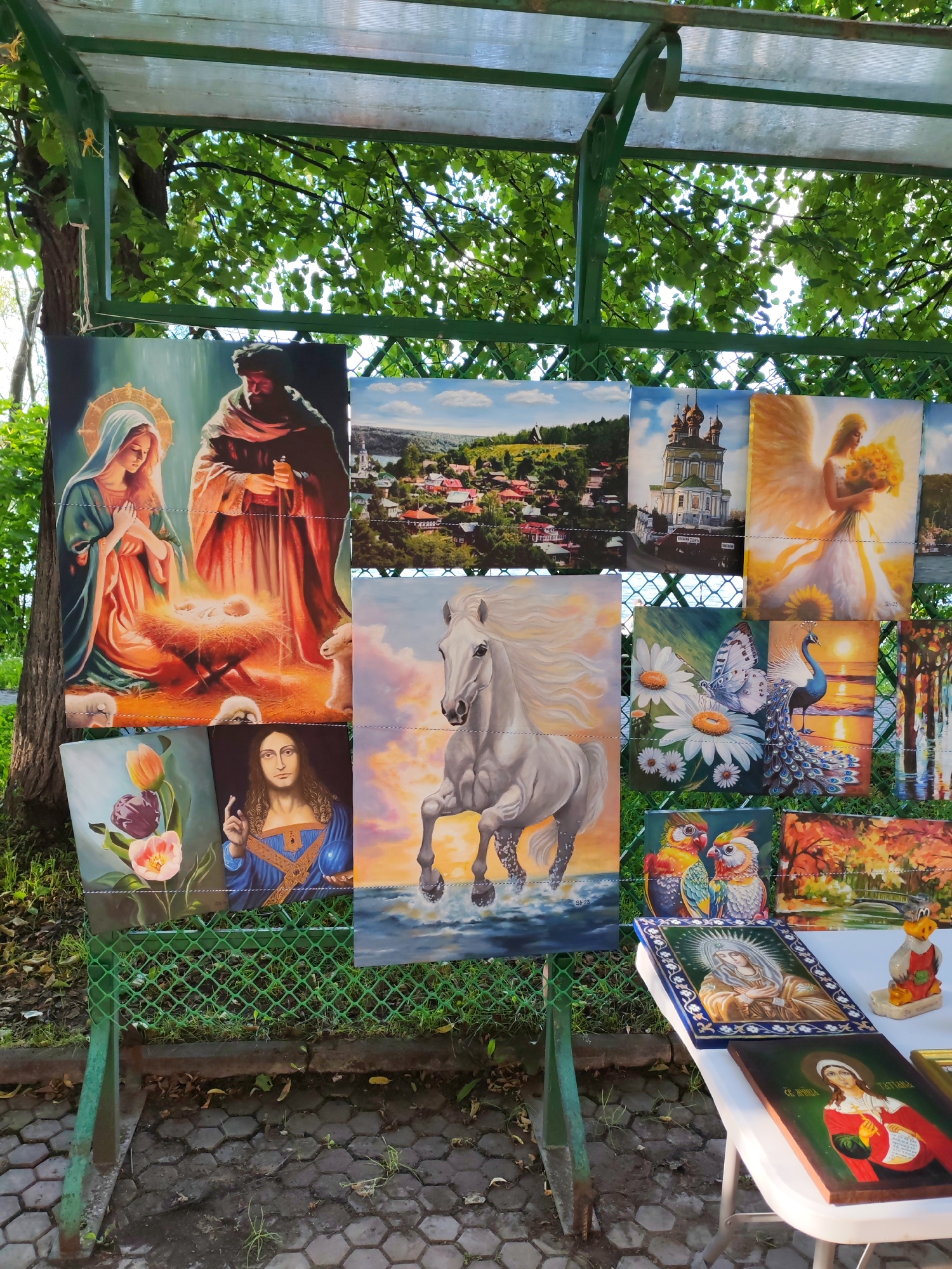 This media file displays a vibrant outdoor art exhibition, likely a market or fair, held in Plyos, Russia. The scene is set under a green metal awning with a translucent roof, offering shade from the elements, while lush green trees provide a natural backdrop. Sunlight filters through the leaves, suggesting a bright, possibly daytime, scene with pleasant weather.
The exhibition features a diverse collection of paintings and a few decorative objects. Religious iconography is prominent, with depictions of the Virgin Mary and baby Jesus, and a painting of Christ. There are also paintings of an angel holding sunflowers, a detailed landscape of a Russian town with its iconic churches, and a striking image of a white horse in motion. Nature is also a recurring theme, showcased in paintings of tulips, a butterfly and peacock by a sunset, and a colorful autumnal landscape. Two colorful parrots add a touch of whimsy to the display.
On a white table in the foreground, several framed religious icons are placed, including one of the Virgin Mary and another with Cyrillic text that reads "СВ. ТАТЬЯНА" (St. Tatiana). A small, whimsical duck figurine also rests on the table, adding to the eclectic nature of the display. The ground is paved with hexagonal stones, typical of public pathways or market areas. The overall impression is one of a lively, creative atmosphere where local art is presented to the public.