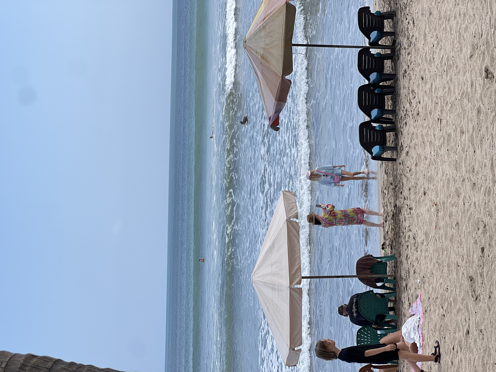 The image depicts a crowded beach scene in Kuta, Indonesia. The foreground is dominated by a sandy beach with several beach chairs and two large umbrellas providing shade. Several people are visible in the image, including individuals sitting on chairs, walking on the sand, and wading in the water. In the background, the ocean stretches out with moderate waves, and a few surfers can be seen in the water. The sky is overcast, suggesting a typical beach day in the region. The scene conveys a sense of leisure and recreation, with a mix of local residents and tourists enjoying the coastal environment.