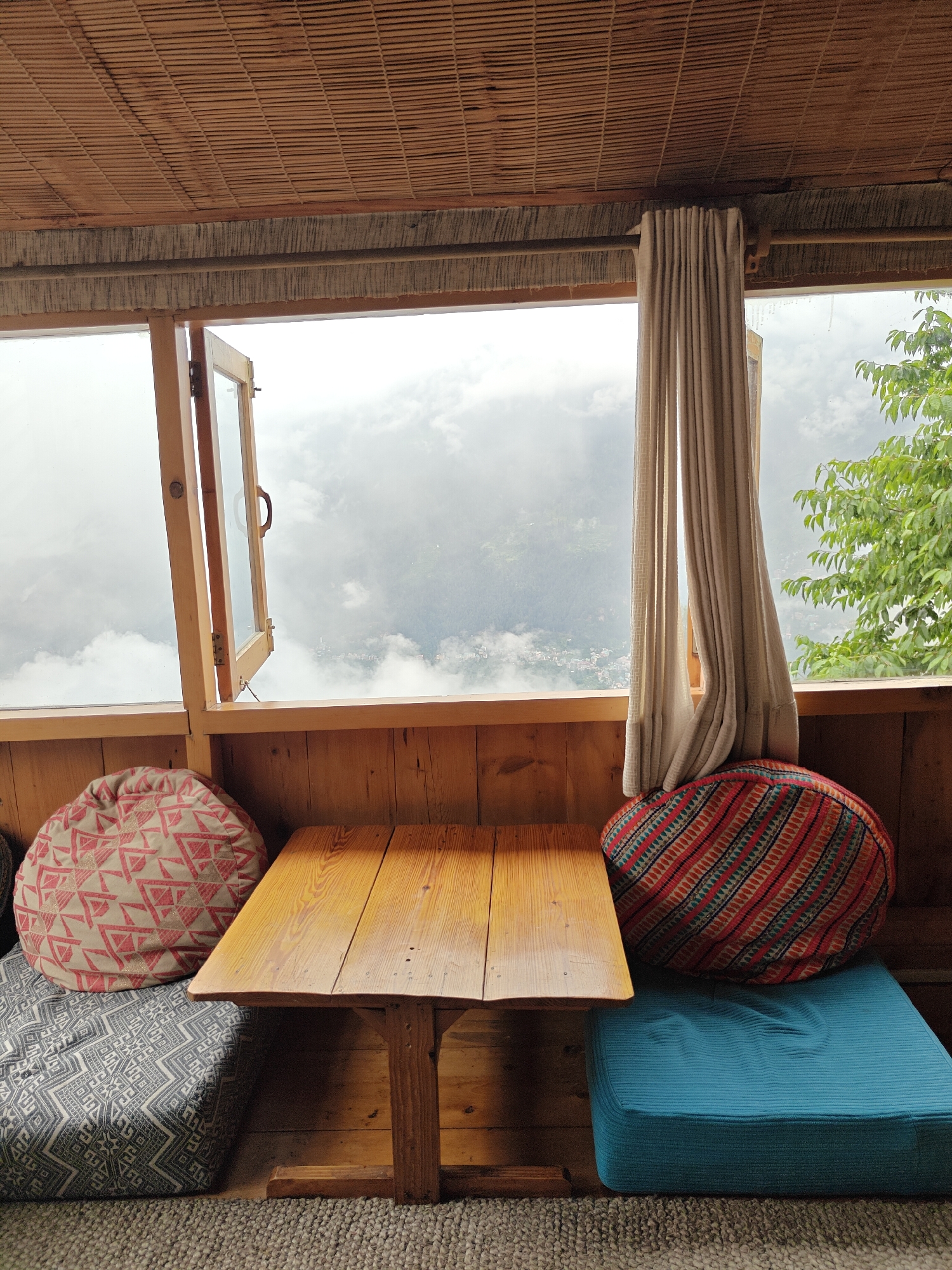 The image depicts an indoor seating area overlooking a misty mountain landscape. A wooden table with a rustic finish is positioned in the center, flanked by floor cushions. On the left, a round cushion with a pink geometric pattern rests on a larger rectangular cushion featuring a grey and white tribal design. To the right of the table, a round cushion with an orange, red, and blue striped pattern sits atop a bright blue rectangular cushion. The seating area is enclosed by wooden paneling, and a bamboo-slatted ceiling is visible overhead. A partially opened wooden-framed window reveals a panoramic view of fog-shrouded mountains and a distant village. A light-colored curtain hangs on the right side of the window. The overall impression is a cozy, rustic retreat with a stunning natural backdrop, likely in or near the city of Prini, India, known for its mountainous terrain and misty weather.