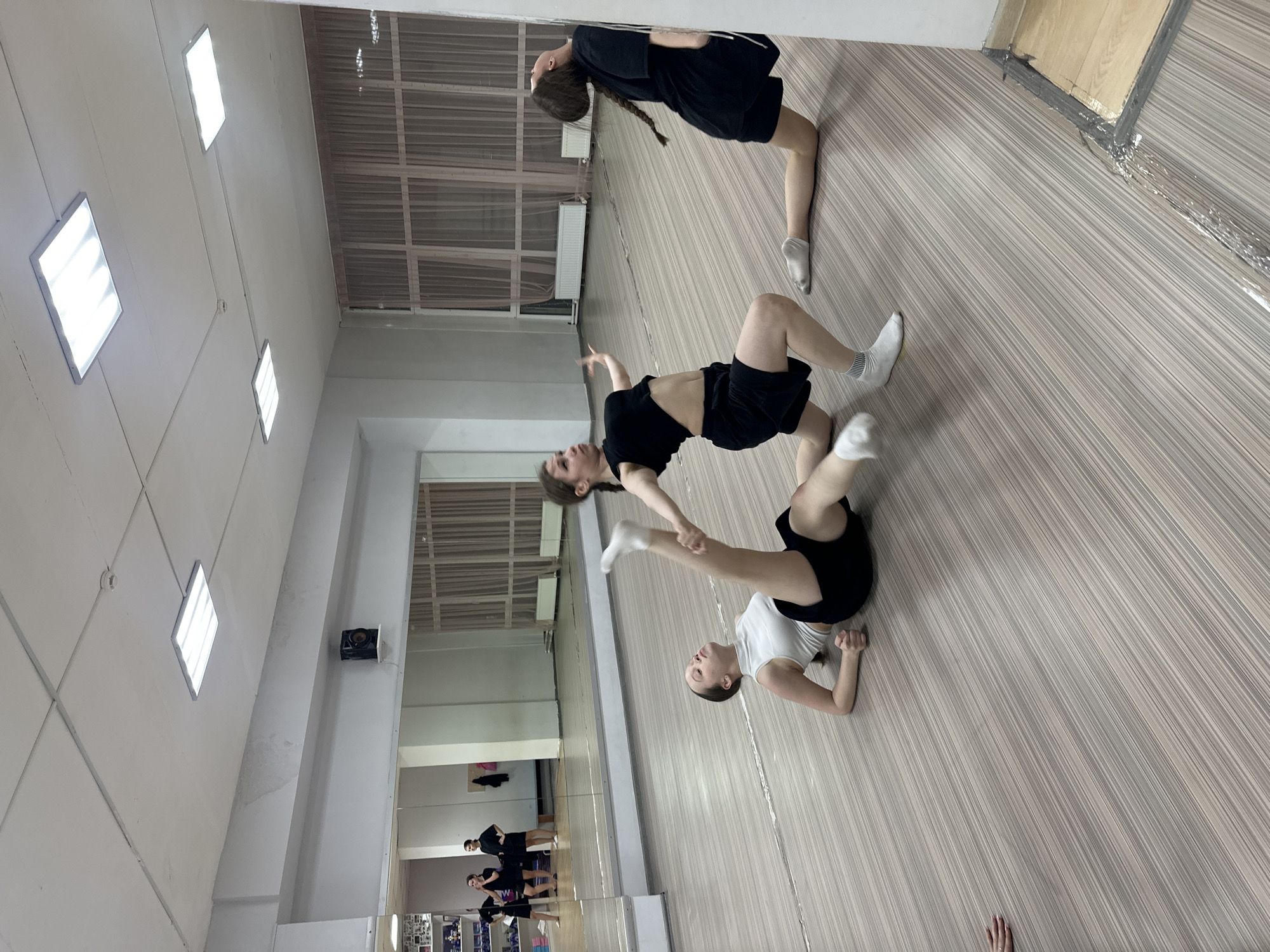 The image captures three women performing a choreographed routine in what appears to be a dance studio or gymnasium. The room is brightly lit with multiple ceiling lights, and the walls are white. A large mirror spans one side of the room, reflecting the dancers and the studio. The floor has a wood-like pattern.
The three women are dressed in athletic wear: one in black shorts and a black top, another in black shorts and a white top, and the third in a black dress or tunic. The women are intertwined, with one woman lying on the floor supporting the legs of another, while the third woman is in a bent-over position.
There is nothing in the image to indicate it is Votkinsk, Russia.