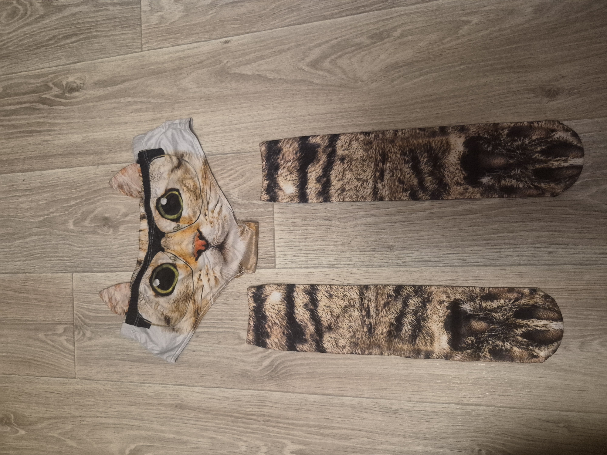 The image shows a pair of cat-themed socks and a garment, likely underwear, printed with a cat's face wearing glasses, laid out on a wood-patterned surface. The location is Unknown, Unknown.