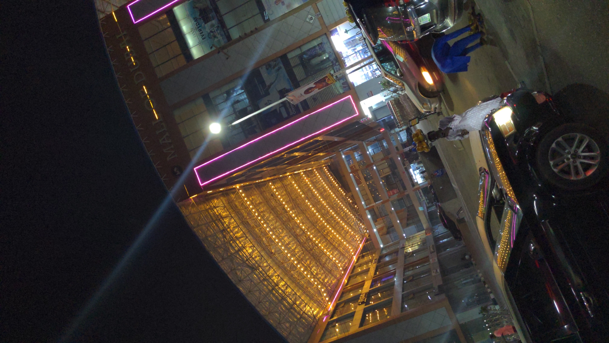 This night-time image, when oriented upright, depicts a bustling commercial scene dominated by a brightly lit, modern multi-story building on the left. The building features a prominent glass facade and is extensively illuminated with warm golden lights, particularly a large, possibly triangular or pyramidal section. Horizontal magenta neon-like light strips further adorn the upper levels.
Visible text on the building includes the word "MALL" on a vertical banner, followed by partially visible letters that seem to spell "DORH" or "DURH," possibly part of a larger name. Another sign on an upper floor clearly reads "DAYS EVERYDAY."
On the right side of the frame, several vehicles are parked along what appears to be a street or mall entrance. A dark-colored SUV or car is prominent in the foreground, with its headlights on and decorative string lights visible on its exterior. Another lighter-colored vehicle, possibly a van or truck, is further down the street. Several indistinct figures, including one person in blue clothing near a silver vehicle and another in light-colored attire, can be seen in the vicinity of the cars and building entrance.
The time of day is clearly night, evidenced by the extensive artificial lighting. Reflections of the bright lights are visible on the building's glass and the vehicles. The scene conveys a lively, urban atmosphere within a commercial area, likely a shopping mall, in Galadimawa Village, Nigeria.
