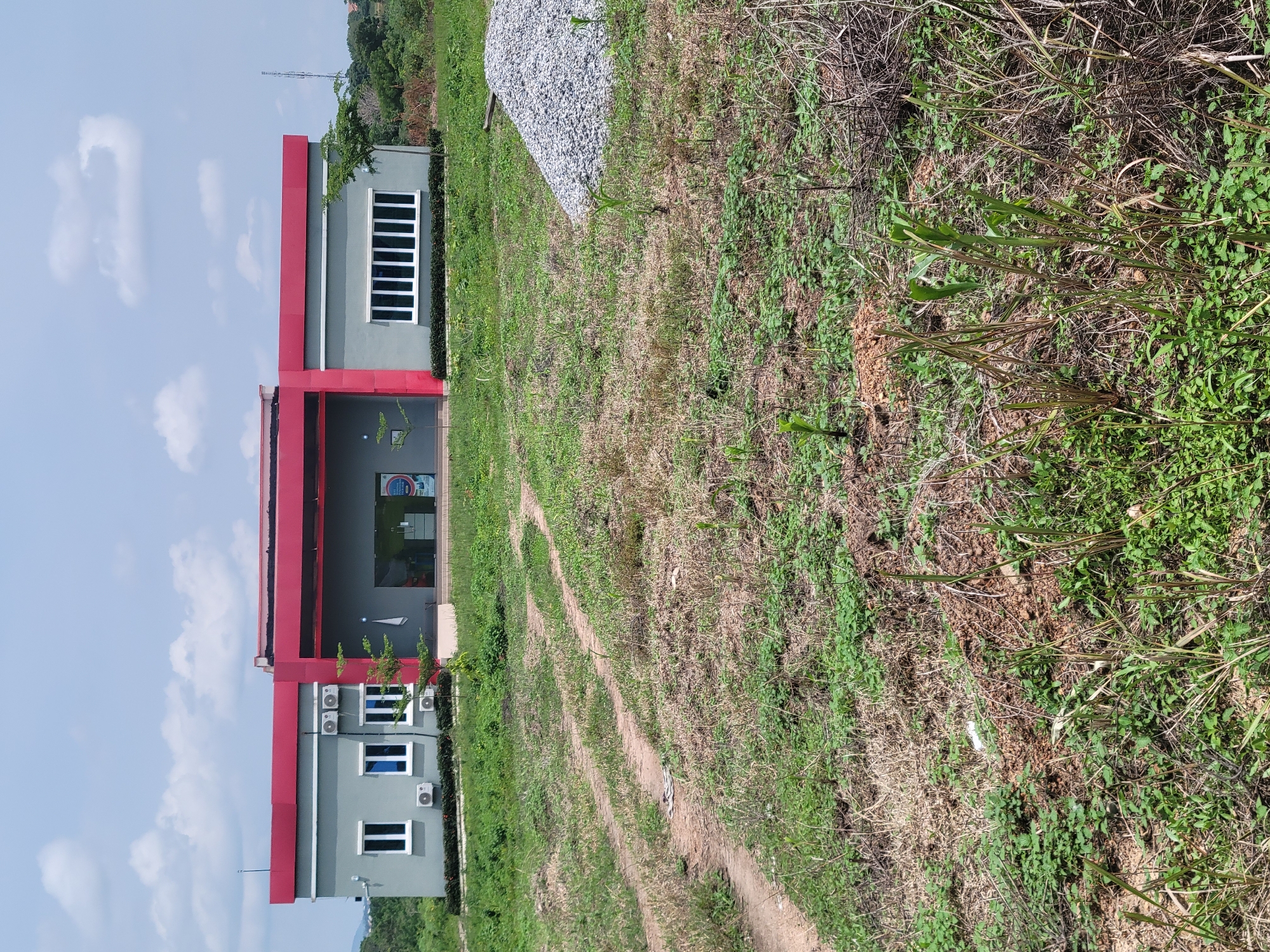 The image depicts a modern, two-story building with a gray facade and vibrant red trim. The building has a flat roof and features several windows, some of which are tinted blue and others with white frames. Multiple air conditioning units are mounted on the exterior walls. The entrance appears to be an open-air walkway with glass doors. A sign with a circular logo and some text is visible above the entrance.
The building is situated on a grassy plot of land with a dirt path winding through it. To the left of the building, there's a stack of gray gravel. The surrounding vegetation is a mix of green grass and dry, brown stalks, suggesting a natural, possibly rural or developing area. The sky is blue with scattered white clouds, indicating daytime. The overall impression is of a newly constructed or under-construction facility in an outdoor setting. No people are visible in the image. The orientation of the image is such that the building appears to be on the left, and the field is on the right, with the sky at the top.