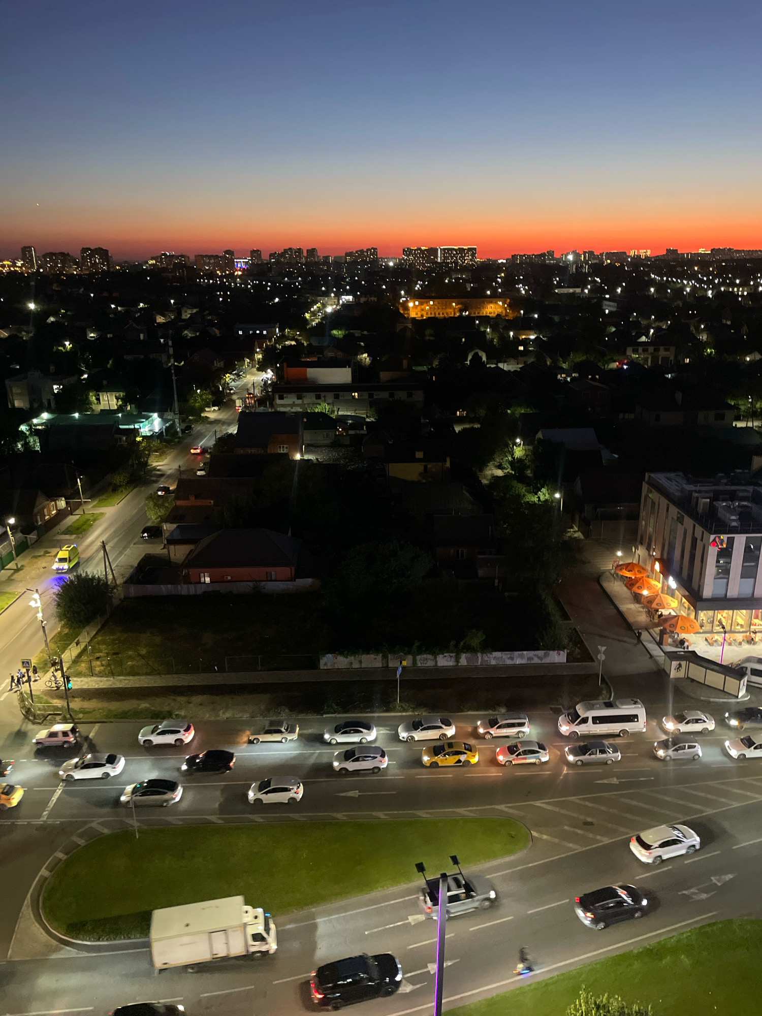 The image captures a panoramic view of a city at dusk, with a vibrant orange and blue gradient in the sky. The scene appears to be in Krasnodar, Russia, as evidenced by the presence of a specific architectural style and city layout typical of the region. The foreground features a multi-lane road with several vehicles, including cars and a white van. Traffic is moderately heavy, with cars moving in various directions. Some cars are parked along the roadside, and a few pedestrians are visible on the sidewalks.
The middle ground shows a mix of residential and commercial buildings. Many houses have illuminated windows, suggesting an evening scene. There are also streetlights and lights emanating from businesses, creating a warm glow across the urban landscape. The background is dominated by a cityscape with taller buildings, likely apartments or office blocks, also lit up against the darkening sky. The overall impression is one of a bustling city transitioning from day to night, with ongoing urban activity. The image is taken from an elevated perspective, offering a broad view of the city's sprawl.