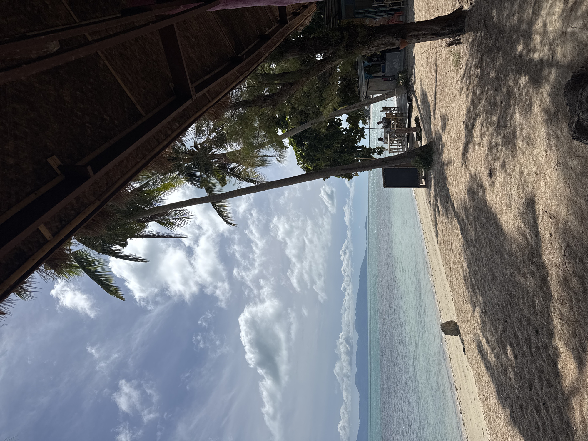 This image captures a serene beach scene in Baan Tai, Thailand. The foreground is dominated by a sandy beach, dappled with the shadows of palm trees that arch over the scene from the right. A rustic, thatched-roof structure, likely a beach hut or restaurant, extends into the frame from the right side. In the mid-ground, the turquoise ocean stretches out to meet a distant horizon line marked by a hazy mountain range. Scattered along the beach are a few objects, including a dark, rectangular signpost and a small, rough-hewn rock. A few people are visible in the distance, seated at tables on a wooden deck, suggesting a casual dining or relaxation spot. The sky above is a blend of blue with scattered white clouds, hinting at a bright, sunny day. The overall impression is one of tropical tranquility and a relaxed atmosphere.