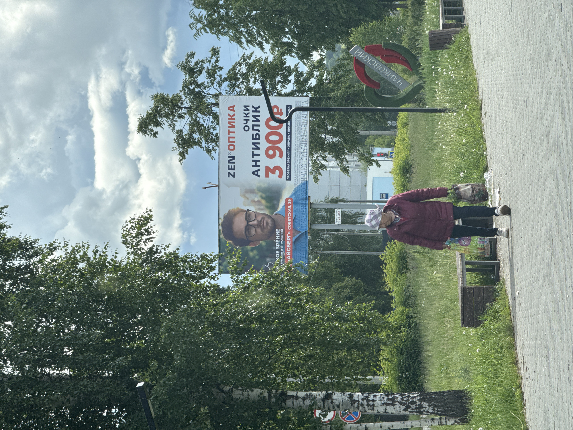 A daytime outdoor photograph, taken in Chaikovsky, Russia, shows a street scene with an older woman walking along a paved sidewalk next to a grassy area. The sky is partly cloudy, with bright patches.
The primary subject is an elderly woman, dressed in a maroon jacket, dark trousers, and a plaid headscarf. She carries a patterned tote bag in her right hand and appears to be walking away from the camera, looking slightly downwards. To her right, a brown wooden bench is visible in the grassy verge, which is dotted with dandelions. Beyond the grass, there are numerous green trees, suggesting a park-like environment or a tree-lined street.
A large billboard dominates the left side of the frame, advertising an optical store. The billboard features a man wearing glasses and prominent Russian text. The upper part reads "ZEN®ОПТИКА" (ZEN OPTICS) and "очки АНТИБЛИК" (anti-glare glasses), with a price of "3 900 Р" (3900 Rubles). Smaller text indicates "ПРОВЕРКА ЗРЕНИЯ + ОПРАВА + ЛИНЗЫ" (EYE EXAMINATION + FRAME + LENSES). Below the man's image, text like "ЧЕТКОЕ ЗРЕНИЕ ОТ БЛИКОВ И ОТРАЖЕНИЙ" (CLEAR VISION FROM GLARE AND REFLECTIONS) and the address "«АЙСБЕРГ» СОВЕТСКАЯ, 39" ("ICEBERG" SOVIETSKAYA, 39) are visible. Fine print at the bottom advises that "НЕОБХОДИМА КОНСУЛЬТАЦИЯ СПЕЦИАЛИСТА" (CONSULTATION WITH A SPECIALIST IS NECESSARY).
To the right of the billboard, a decorative street sign spells out "ЧАЙКОВСКИЙ" (CHAIKOVSKY) in large, stylized red and green letters. Further in the foreground, birches and other trees line the sidewalk. The image is taken from a slightly elevated perspective, looking down towards the sidewalk.