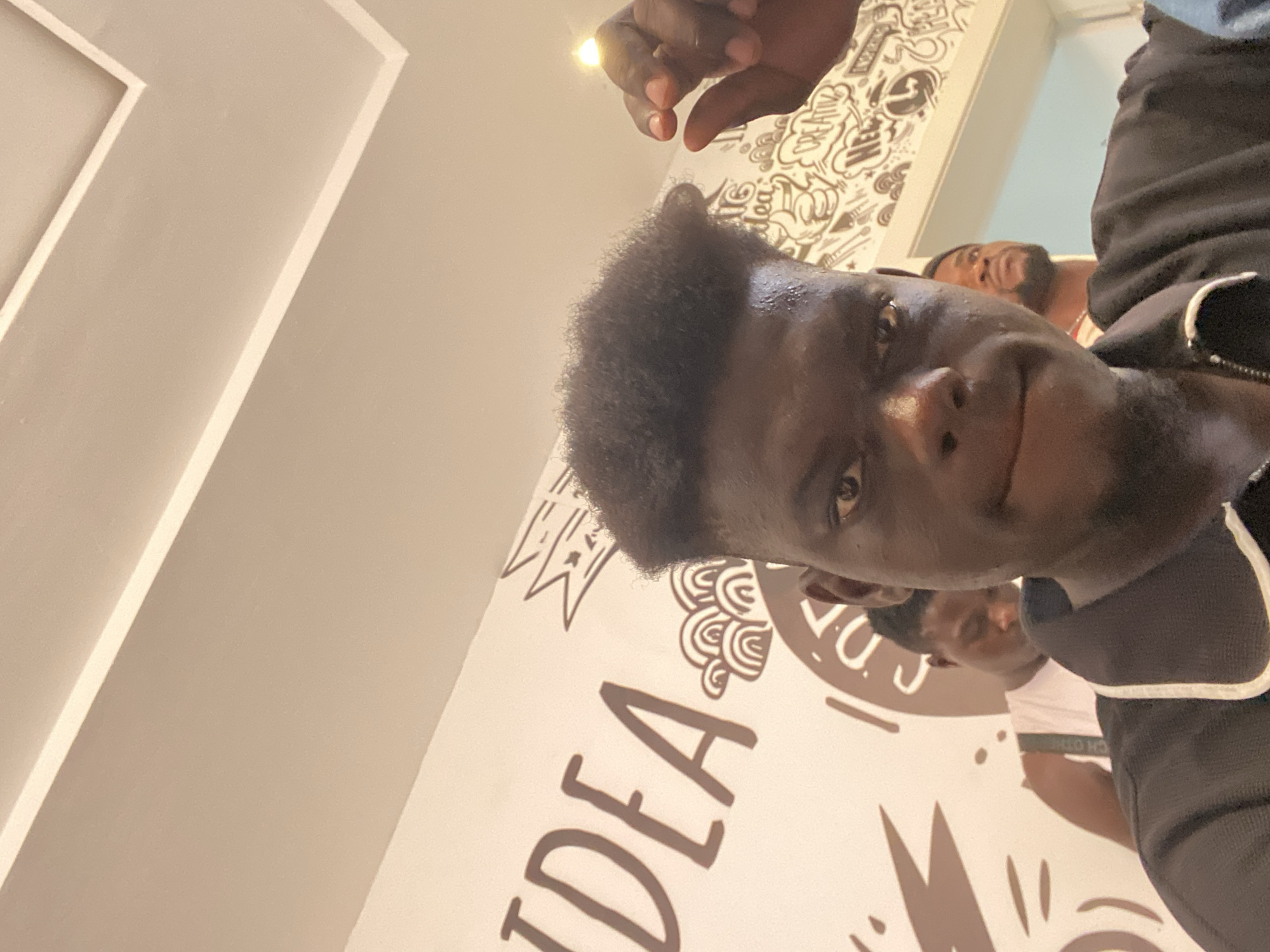 The image is a close-up, low-angle shot of a young Black man looking directly into the camera, giving the impression of a selfie or a candid photo. The man, with short, dark, curly hair, occupies the majority of the foreground, his face well-lit with a bright reflection on his forehead and nose. He is wearing a dark top, possibly a jacket or hoodie, with white trim visible on the collar area. His right hand is visible in the upper part of the frame, near the top edge.
The background features a light-colored wall or ceiling decorated with stylized, doodle-like patterns in a darker color, possibly brown. Prominently displayed on the wall is the large, artistic text "IDEA." Other partial words and decorative elements are scattered across the background, including a partial view of the word "CREATIVE" in one of the crop hints.
Two other individuals are visible in the blurred background. One man with a beard is situated behind the main subject's head, looking upwards. Another person's face is partially visible further back and to the right. The setting appears to be an indoor space, possibly a cafe, creative hub, or office, given the decorative walls. Architectural features like angular lines and molding are also visible on the light-colored surfaces, suggesting structured interior design. The overall lighting is bright, illuminating the scene clearly.