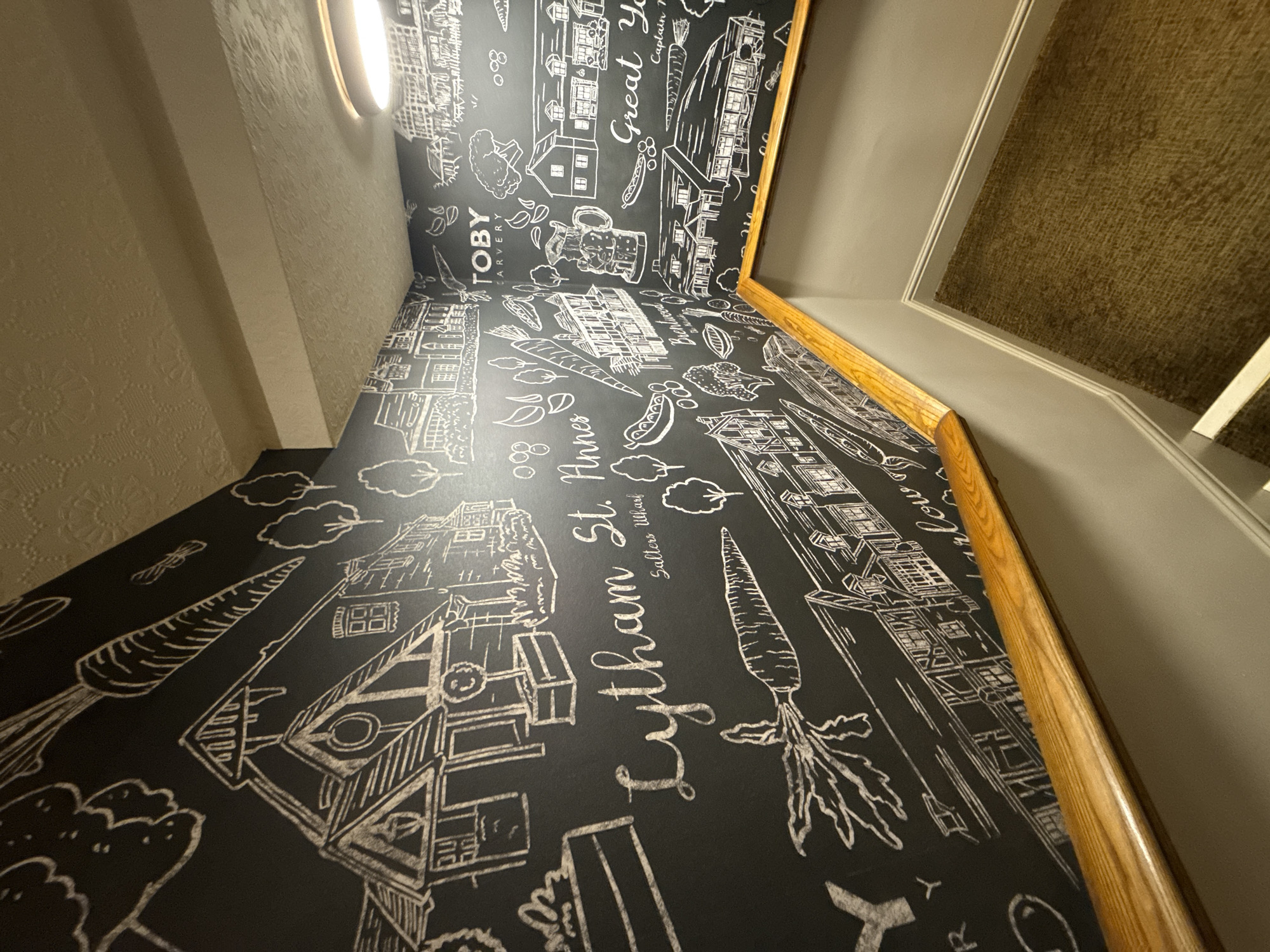 The image is an eye-level, wide-angle shot of a wall with drawings on it. The wall is divided into two sections. The top section is light beige and textured, while the bottom section is black with white line drawings. The drawings appear to be a representation of Lytham St. Annes, a seaside town in Lancashire, England, and Great Yarmouth. The drawings include buildings, carrots, clouds, and writing. The writing includes "Lytham St. Annes," "Salters Wharf," "TOBY CARVERY," and "Great Yarmouth, Captain." A wooden railing runs along the wall, separating the upper and lower sections. There is an elliptical light fixture in the corner of the room. The wall on the right side of the frame is gray with a brown, textured panel in the upper corner.
