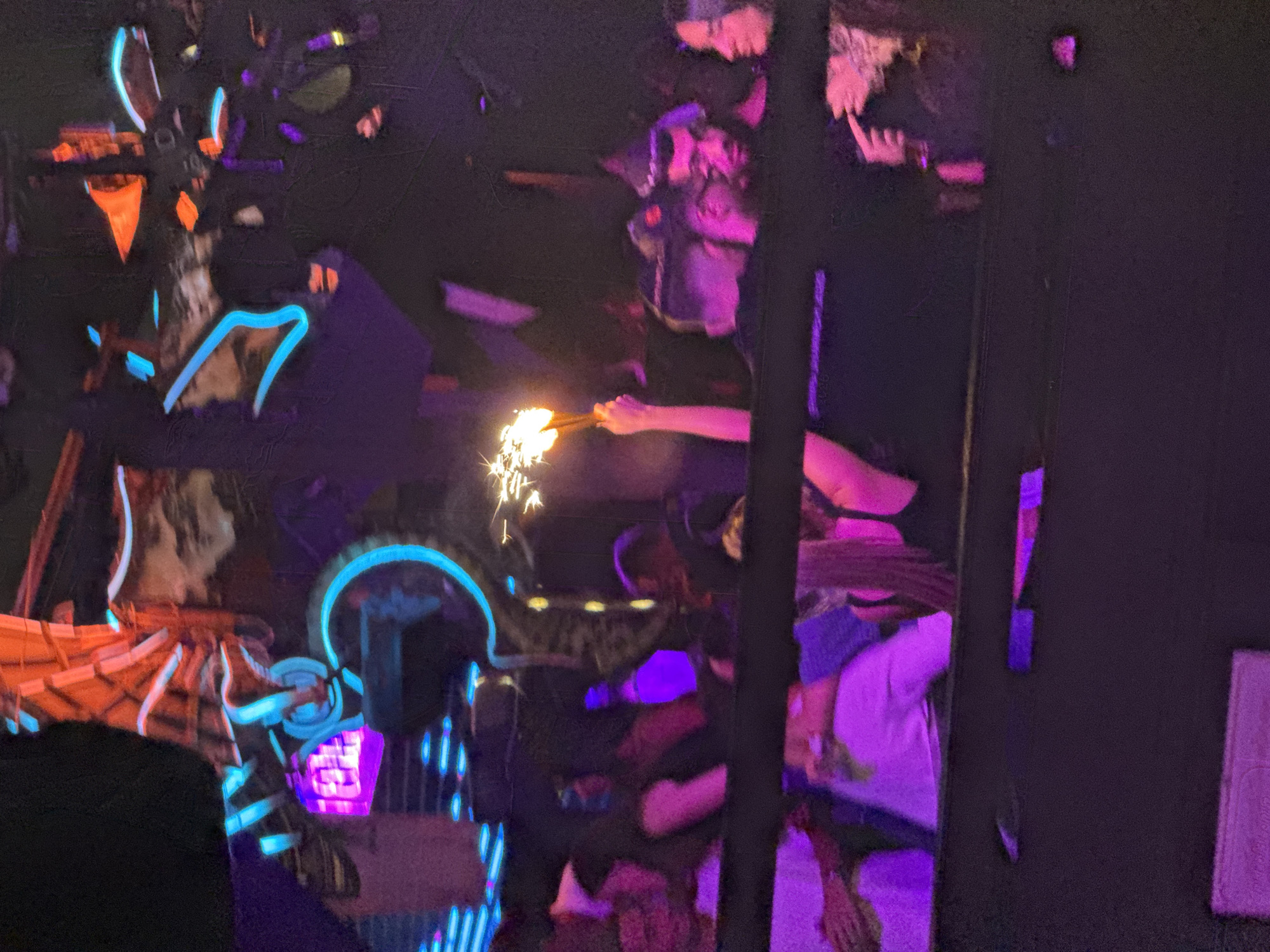 The image appears to depict a scene within a dimly lit environment, possibly a bar or club in Tel-Aviv, Israel, given the location. There are multiple people present, some of whom are holding sparklers, indicating a celebratory occasion. A neon light structure, with elements of blue and orange, is prominently displayed. The overall atmosphere is one of festivity with purple lighting enhancing the mood. The image suggests interactions between individuals with their faces blurred.