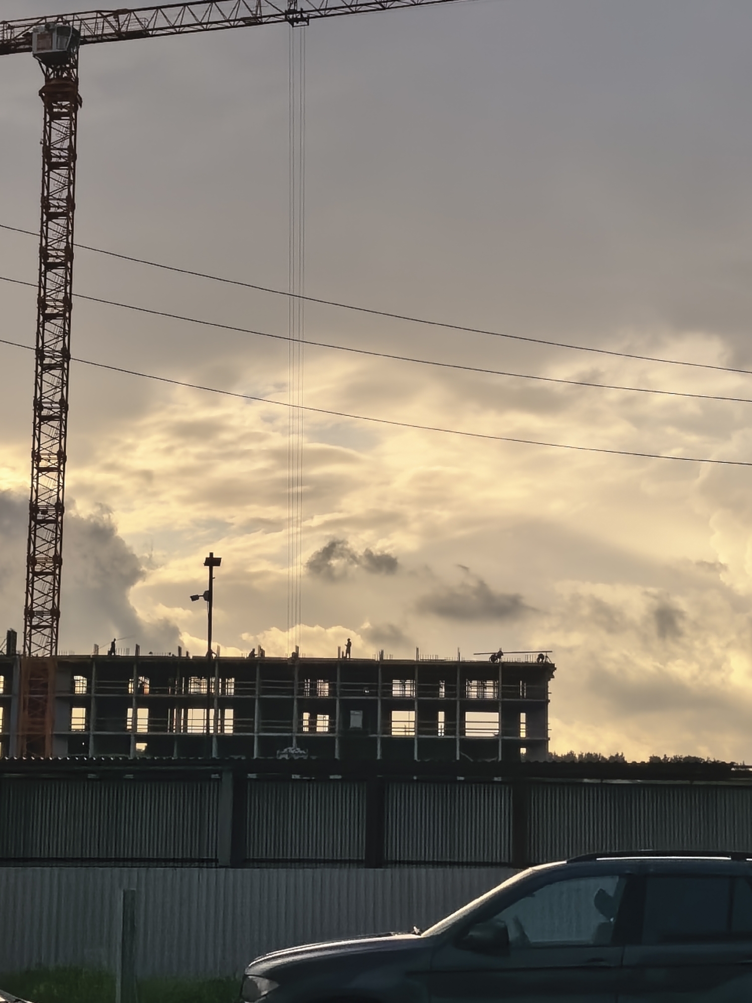 The image captures a construction site during either sunrise or sunset in Unknown, Unknown. A prominent, red-orange tower crane stands on the left, with its lattice structure and cabin visible against the sky. Multiple dark utility lines traverse horizontally across the upper sky, with several vertical cables descending from the crane's boom.
In the midground, a multi-story building under construction is rendered in silhouette against a luminous sky filled with golden-hued and grey clouds. The building exhibits an exposed concrete frame with numerous open window and floor spaces. Approximately six to seven human figures, identified as construction workers, are visible as dark silhouettes on the topmost completed floor of the building, appearing to be standing or moving. One figure on the far right operates a piece of equipment, and a tall, dark, pole-like structure stands near the center of the building's roofline.
A corrugated metal fence, dark grey in color, spans the width of the midground, separating the construction building from the immediate foreground. The lower right foreground displays the partially visible front section of a dark-colored vehicle, likely an SUV, with a small strip of green grass visible below it. The overall lighting is characterized by strong backlighting, creating prominent silhouettes of the structures and figures against the bright, cloud-filled sky.