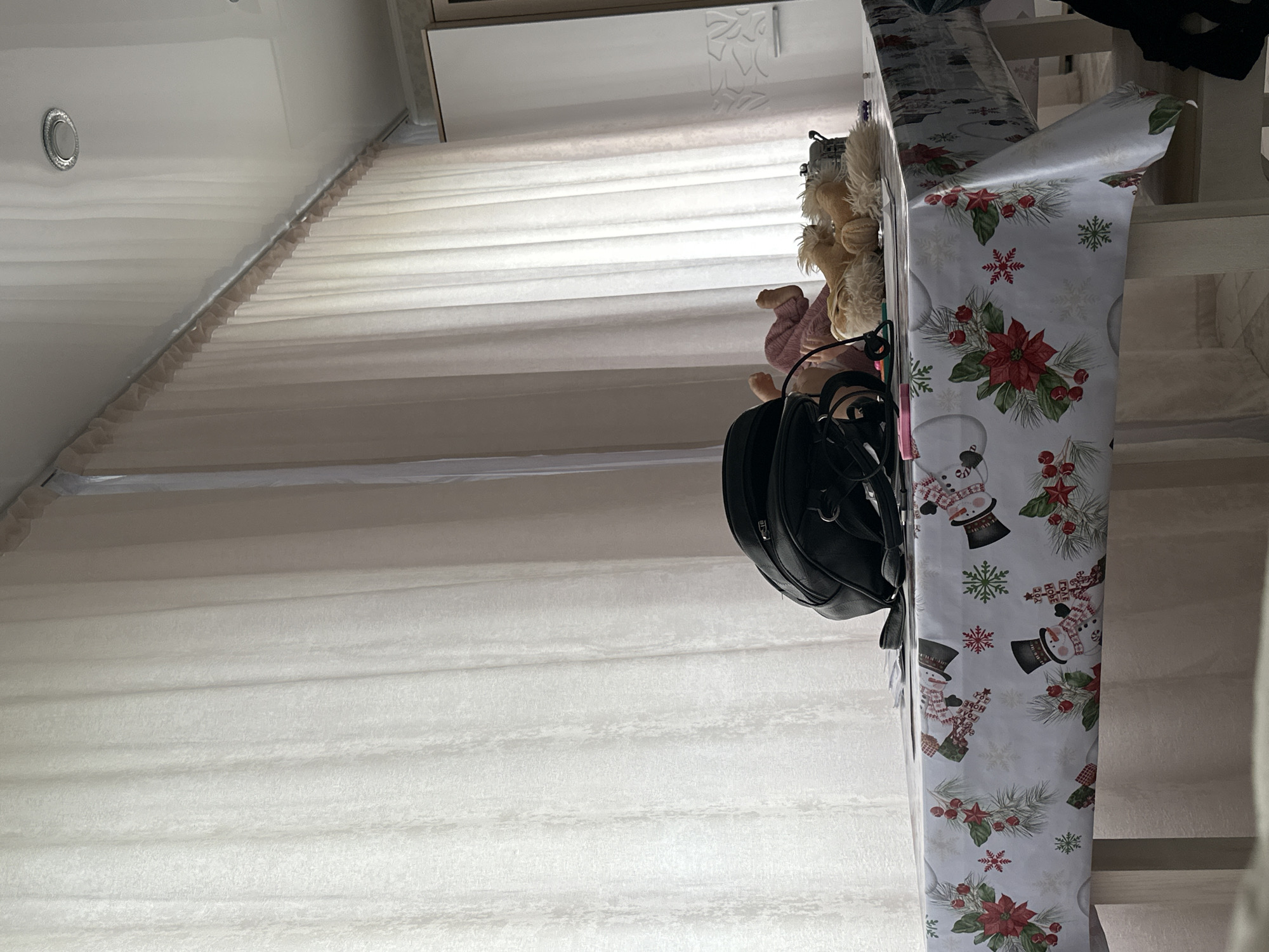 Here is a detailed description of the image:
The image appears to be taken inside a residence, likely a house, given the architectural details. The walls and ceiling are predominantly white, with a subtle striped pattern running horizontally across the frame. The most prominent element is a set of stairs, also white, extending towards a top landing.
On the right side of the frame, partially obscuring the view of the stairs, is a roll of Christmas-themed wrapping paper, featuring images of snowmen, snowflakes, and poinsettias. A black backpack or purse hangs near the wrapping paper. Additionally, a couple of stuffed animals are visible near the wrapping paper.
The environment appears to be clean and well-maintained. The image gives the impression of a cozy and festive atmosphere, presumably during the holiday season.
Without any explicit information, it's impossible to determine the exact location in terms of the city. Therefore, the city of "Unknown, Unknown" would be the accurate answer.