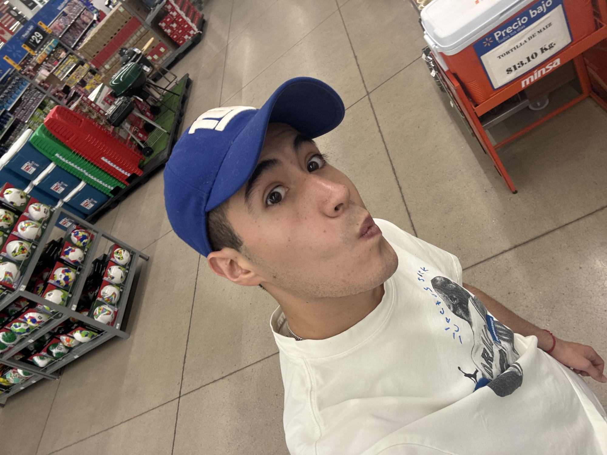 A young man poses for a selfie inside what appears to be a Walmart store in San Bernardino Tlaxcalancingo, Mexico. He is positioned at an upward angle, looking directly at the camera with wide, slightly surprised eyes and puckered lips, indicating a playful or humorous expression. He wears a blue baseball cap with a white "F1" or similar logo, a white t-shirt featuring a graphic print of a shoe (possibly a sneaker from Travis Scott's line, with "JACKS BACK" or similar text visible), and a silver chain around his neck.
The background reveals typical supermarket aisles. To the left, shelves are stocked with various items, including small soccer balls on a multi-tiered display rack. A green and red cooler or storage bin is also visible on a shelf, and further back, more retail shelves with products. A price tag displaying "$29" is visible on one of the shelves. To the right, a red stand holds a white cooler with a sign attached. The sign, featuring a Walmart logo, advertises "Precio bajo" (low price) for "TORTILLA DE MAIZ" (corn tortilla) at "$13.10 Kg." (13.10 pesos per kilogram), and the brand "minsa" is also visible, confirming the product and local context. The flooring is a light-colored tiled surface, commonly found in large retail stores. The lighting suggests an indoor setting, likely during daylight hours given the store environment.